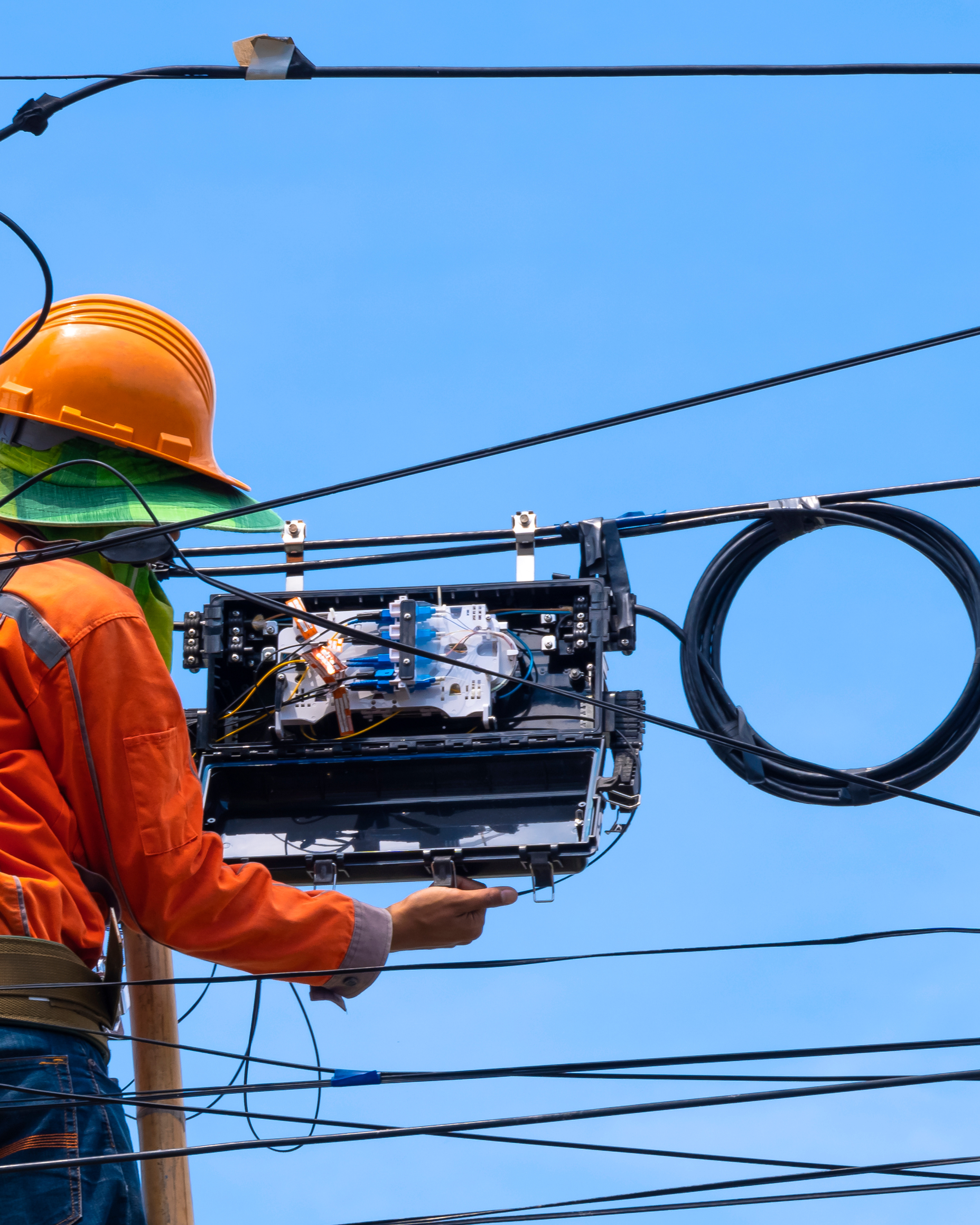 A worker wearing an orange safety vest and yellow hard hat working on electric or telecommunication lines on a utility pole against a clear blue sky.