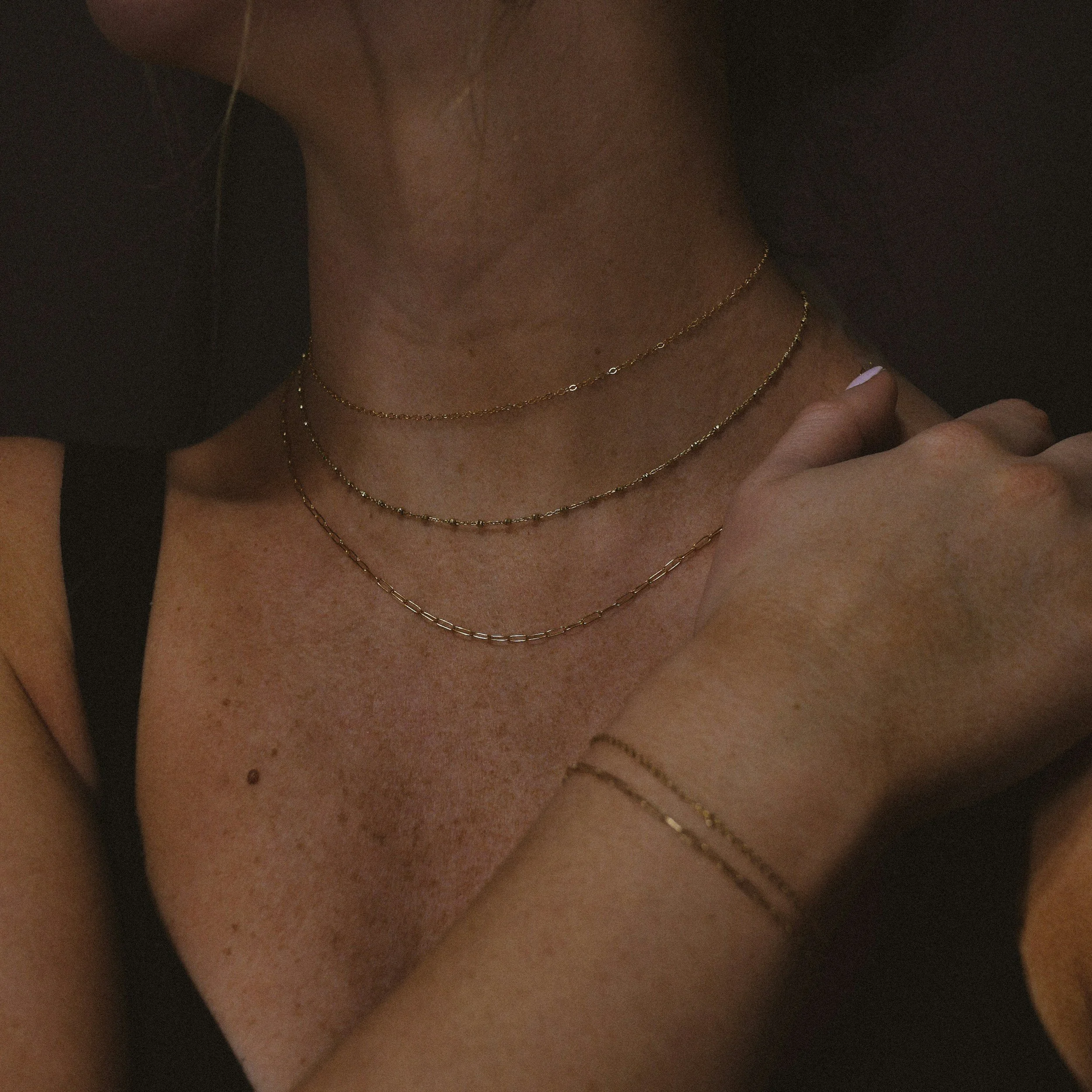 Close-up of a person's neck and chest with layered gold necklaces, holding their collarbone. permanent jewelry in Virginia, DC, Maryland