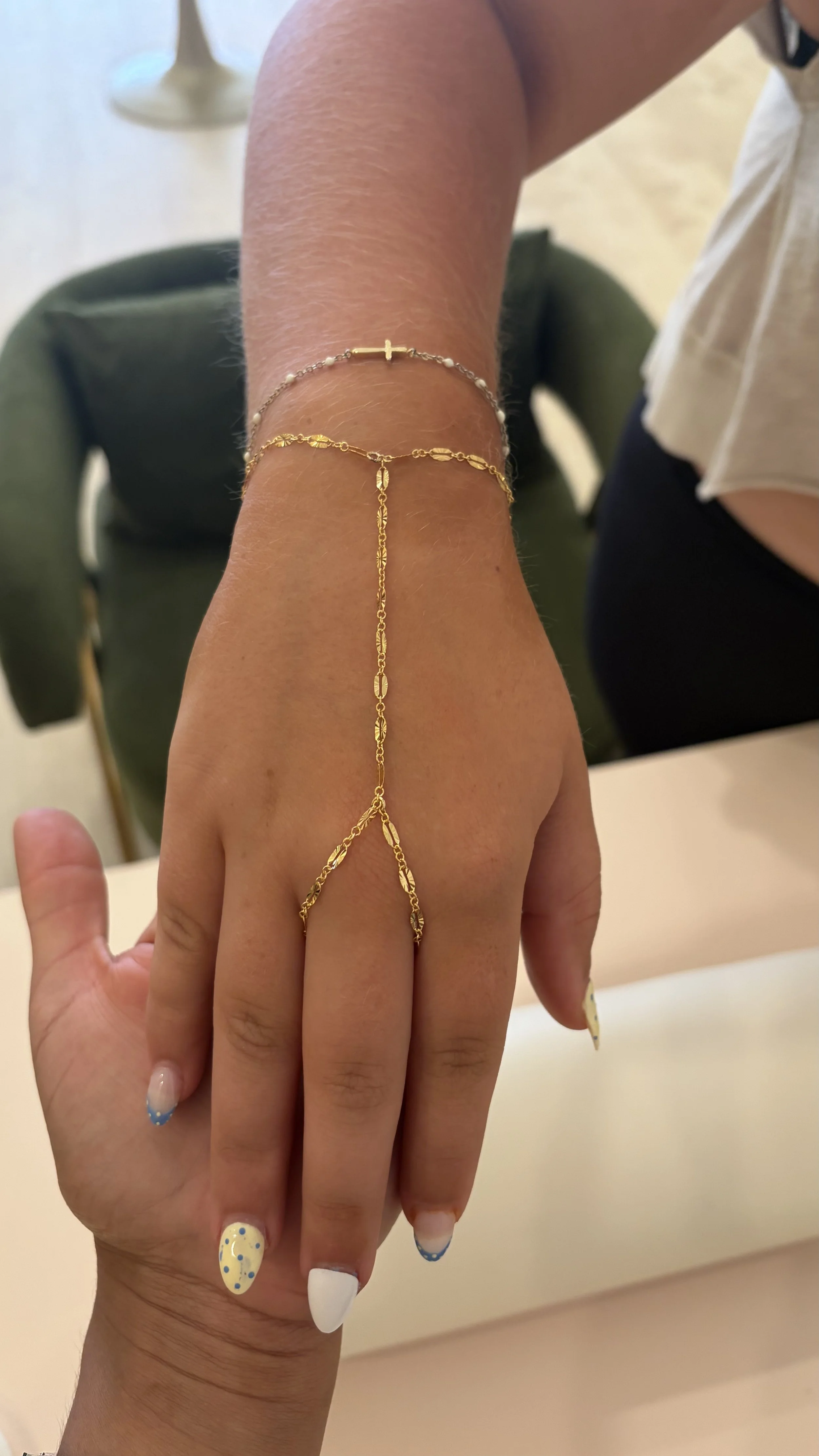 Close-up of a person's hand and wrist, adorned with multiple gold and silver bracelets (hand chain), with colorful nail polish. The background shows part of a table and a chair.permanent jewelry in Virginia, DC, Maryland