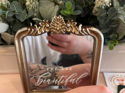 Decorative mirror with gold ornate frame reflecting a person holding a plant, with a sign that says 'You Are Beautiful'.