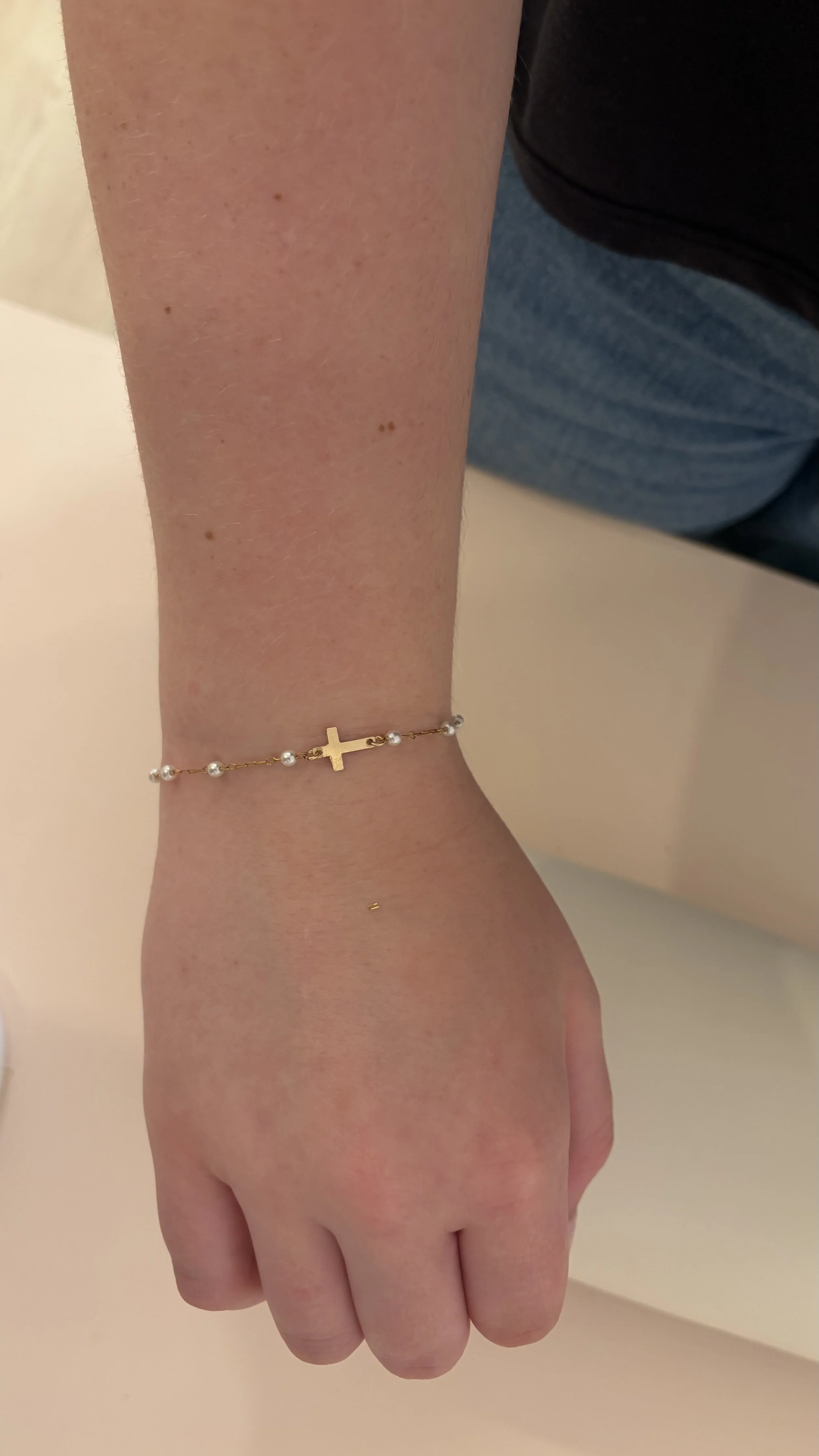 Close-up of a person's wrist wearing a delicate gold bracelet with small pearls and a cross charm. The person has fair skin and is wearing a dark top and blue jeans, with a plain light-colored background. permanent jewelry in Virginia, DC, Maryland 