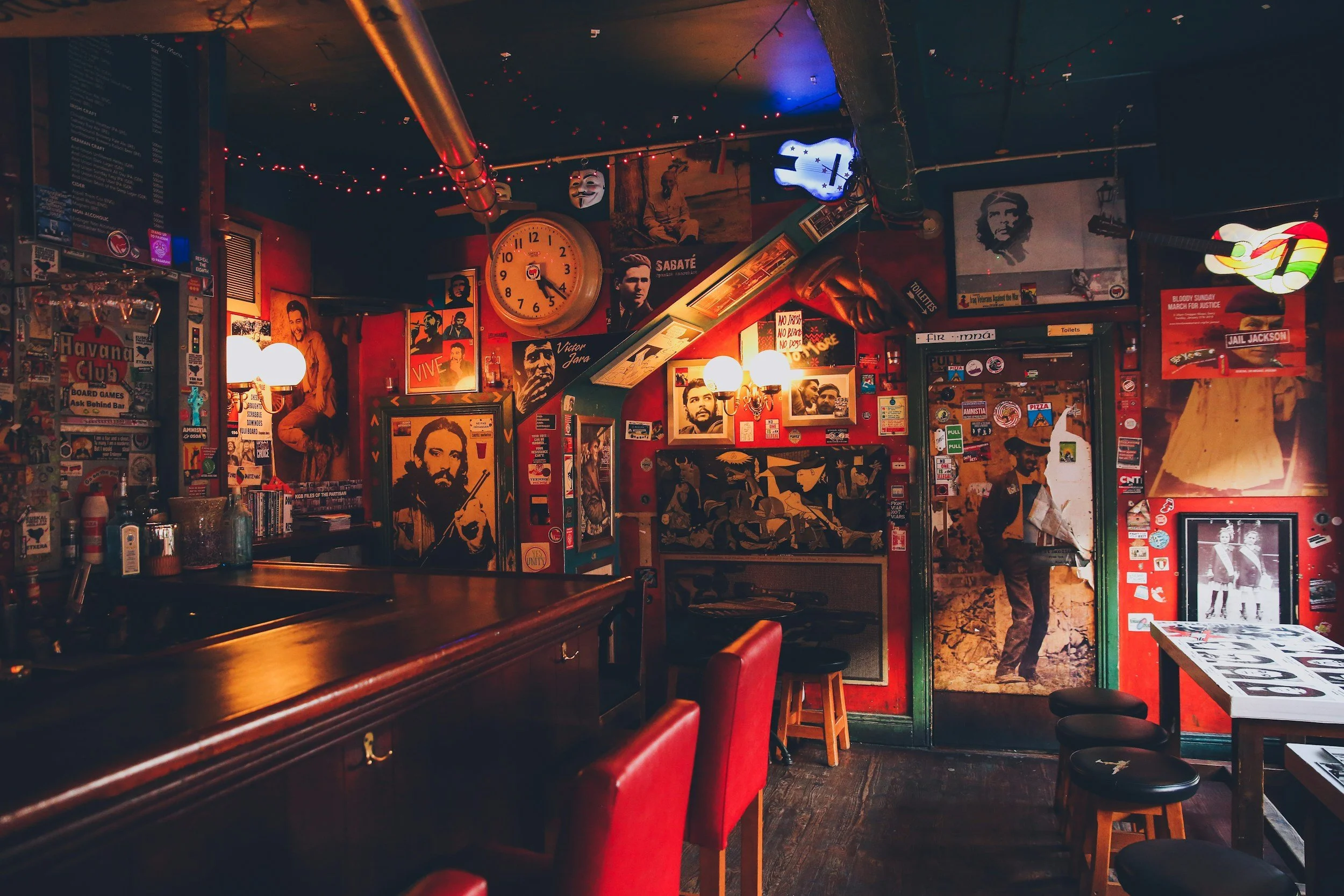 Interior of a cozy bar or cafe with red walls decorated with posters of famous personalities, a large clock, and string lights. There are tables and chairs, including red upholstered chairs at the bar, and vintage artwork and memorabilia on the walls.