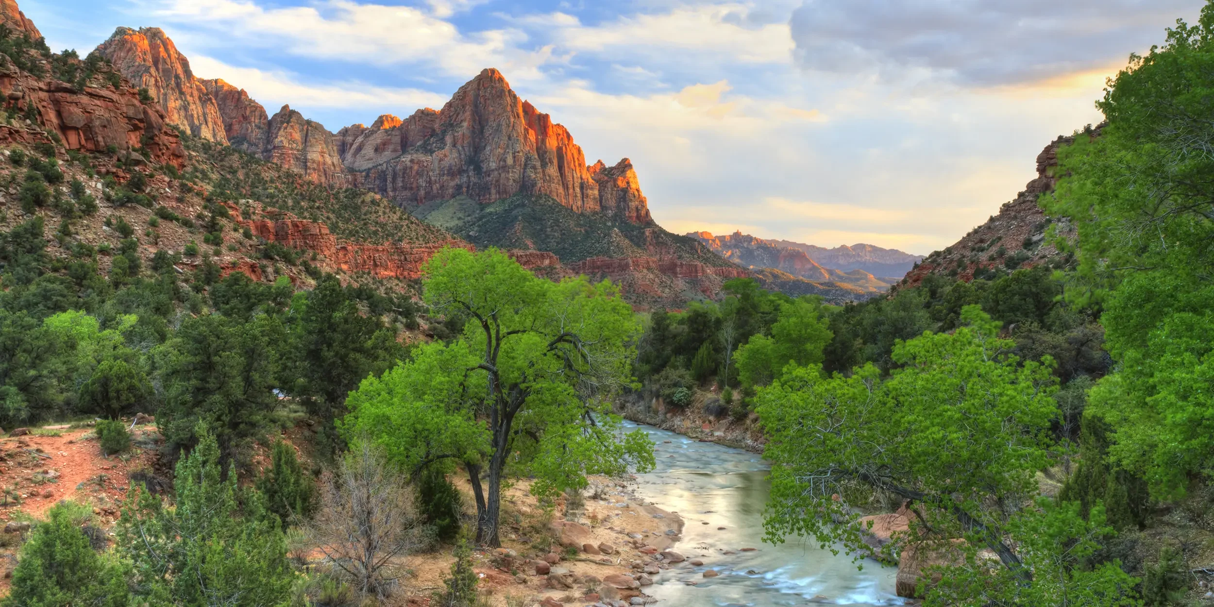 Scenic view of Zion National Park featuring red rock cliffs, green trees, and a river flowing through the valley at sunset.