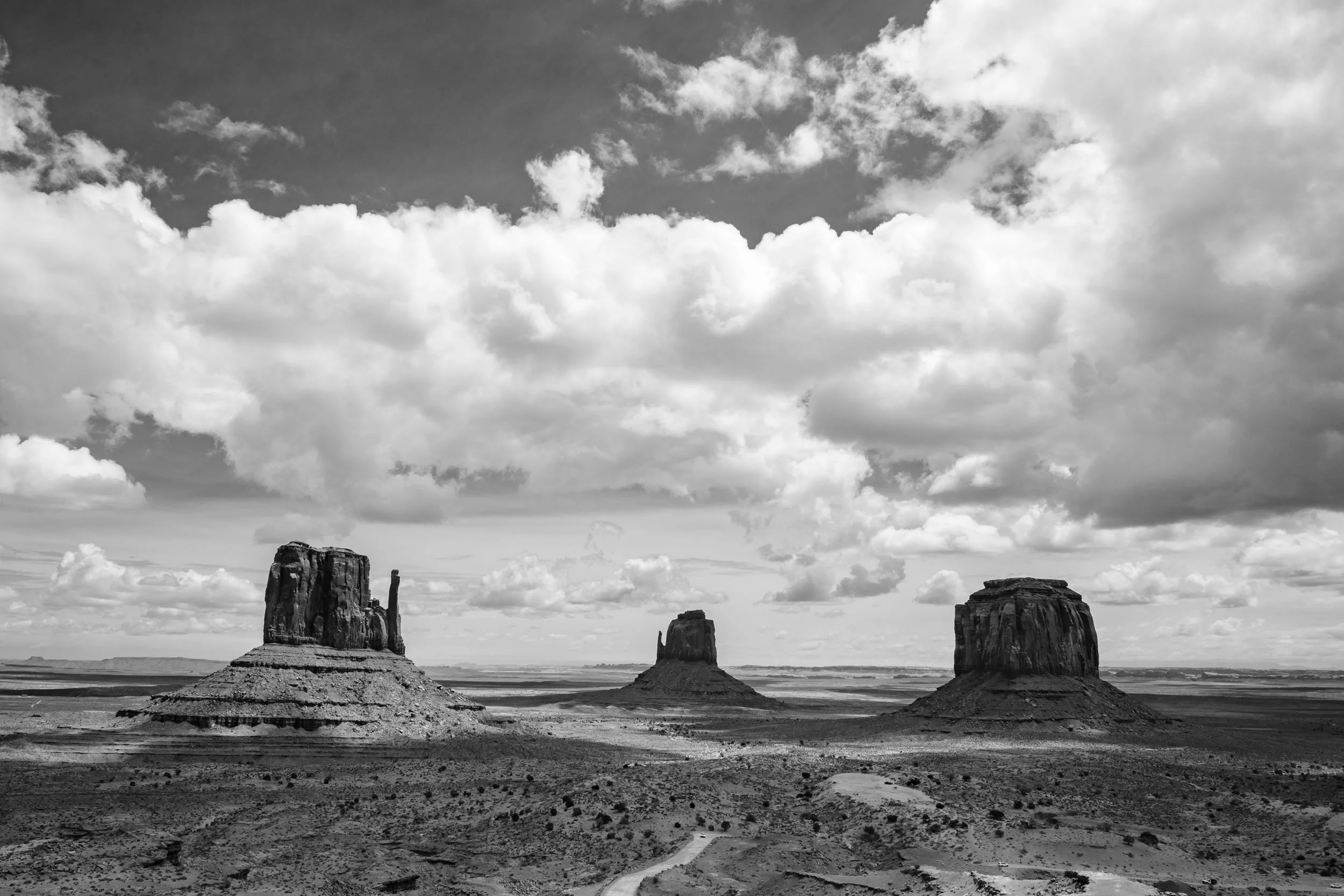 Black and white landscape photo of Monument Valley with prominent buttes and mesas in the distance, under a cloudy sky.