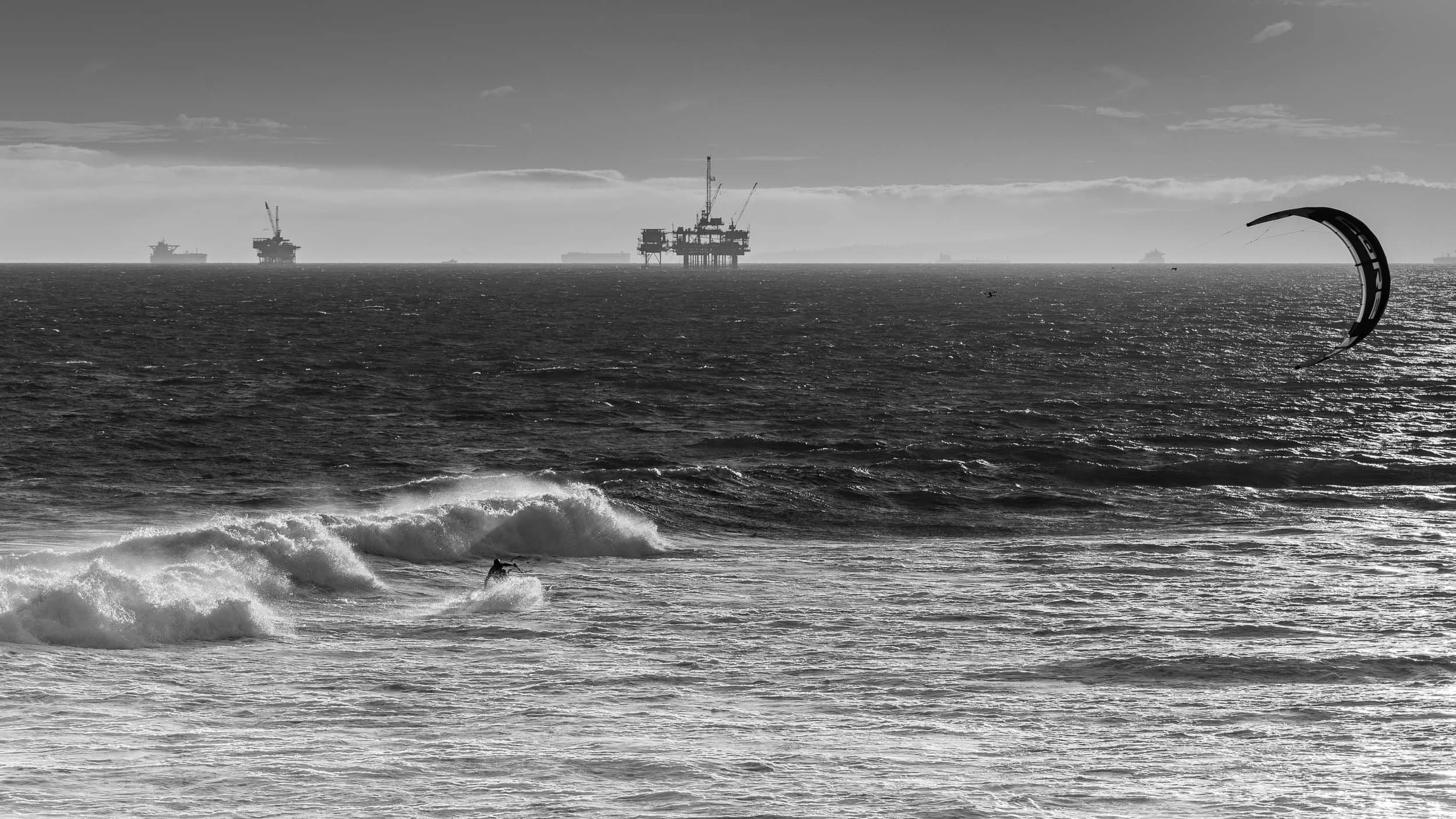 A black and white photo of the ocean with waves and a surfer riding a wave in the foreground. In the background, there are multiple offshore oil rigs and ships on the horizon, and a kite surfer with a curved kite to the right.