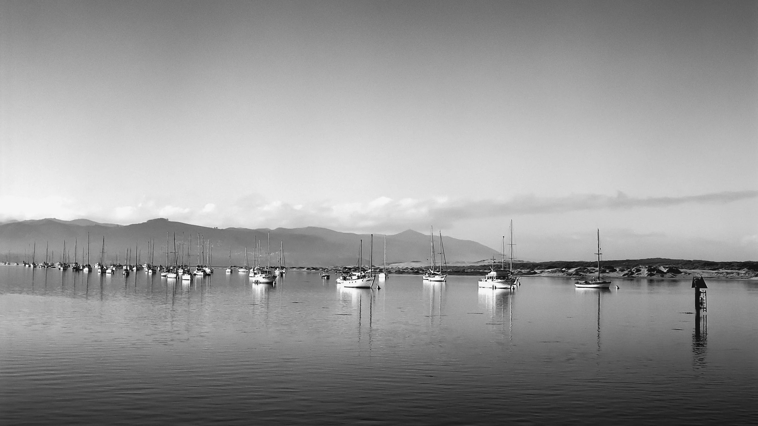 Black and white photo of sailboats anchored in a calm bay with mountains in the background and a clear sky.
