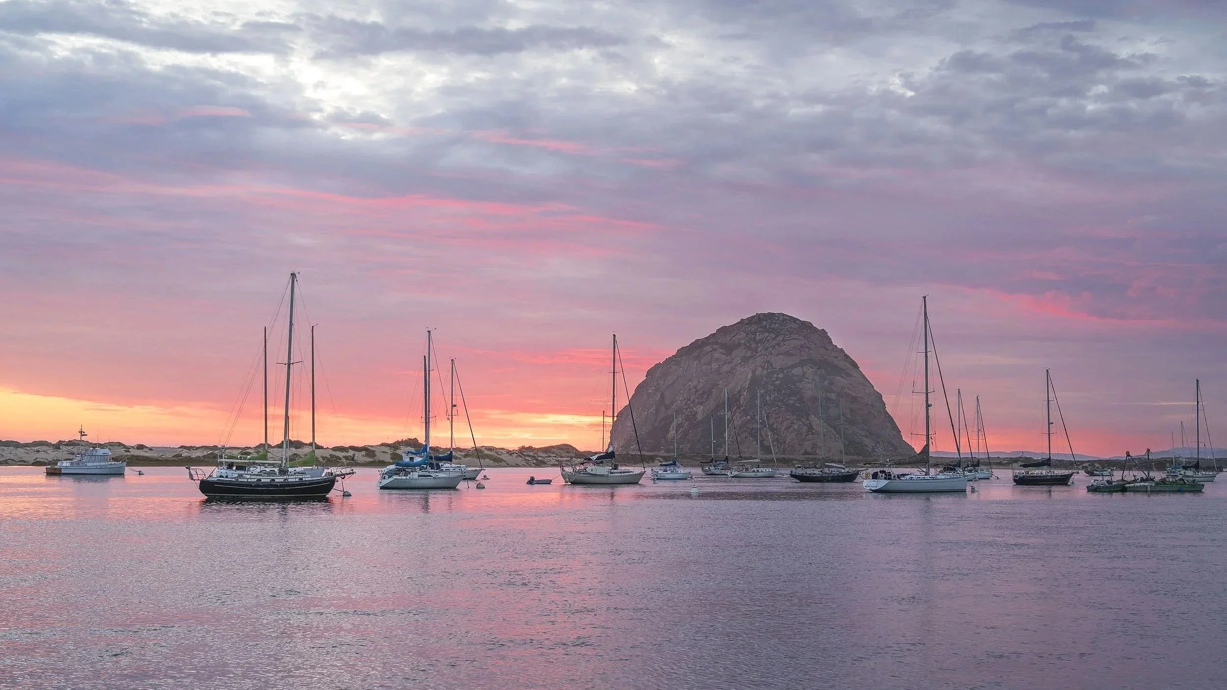 Sailboats anchored in a calm harbor during sunset with a large rock formation and a colorful sky with pink and purple clouds.