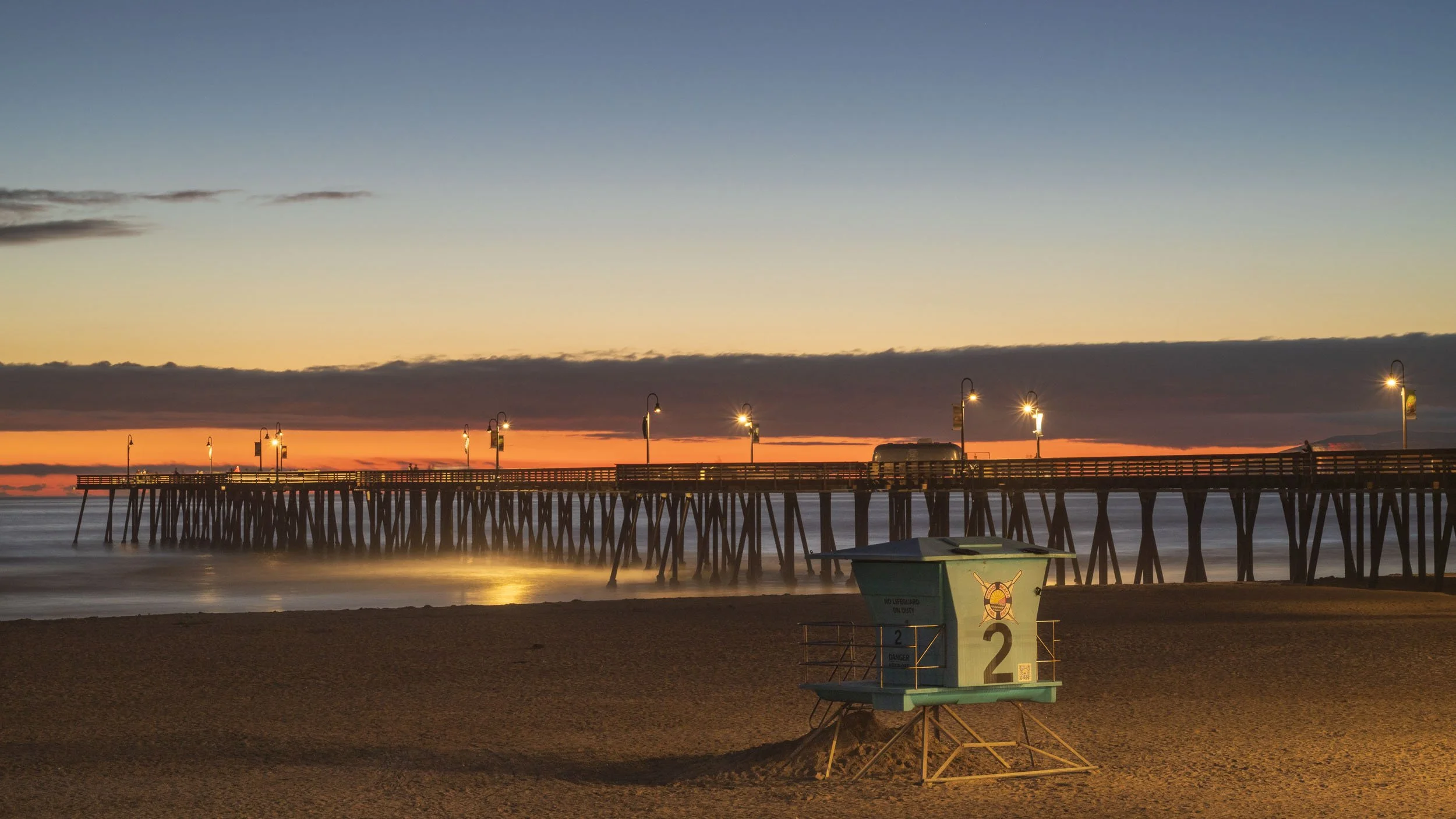 Sunset over a pier with glowing lamps, calm water, and a lifeguard station in the foreground, on a sandy beach.