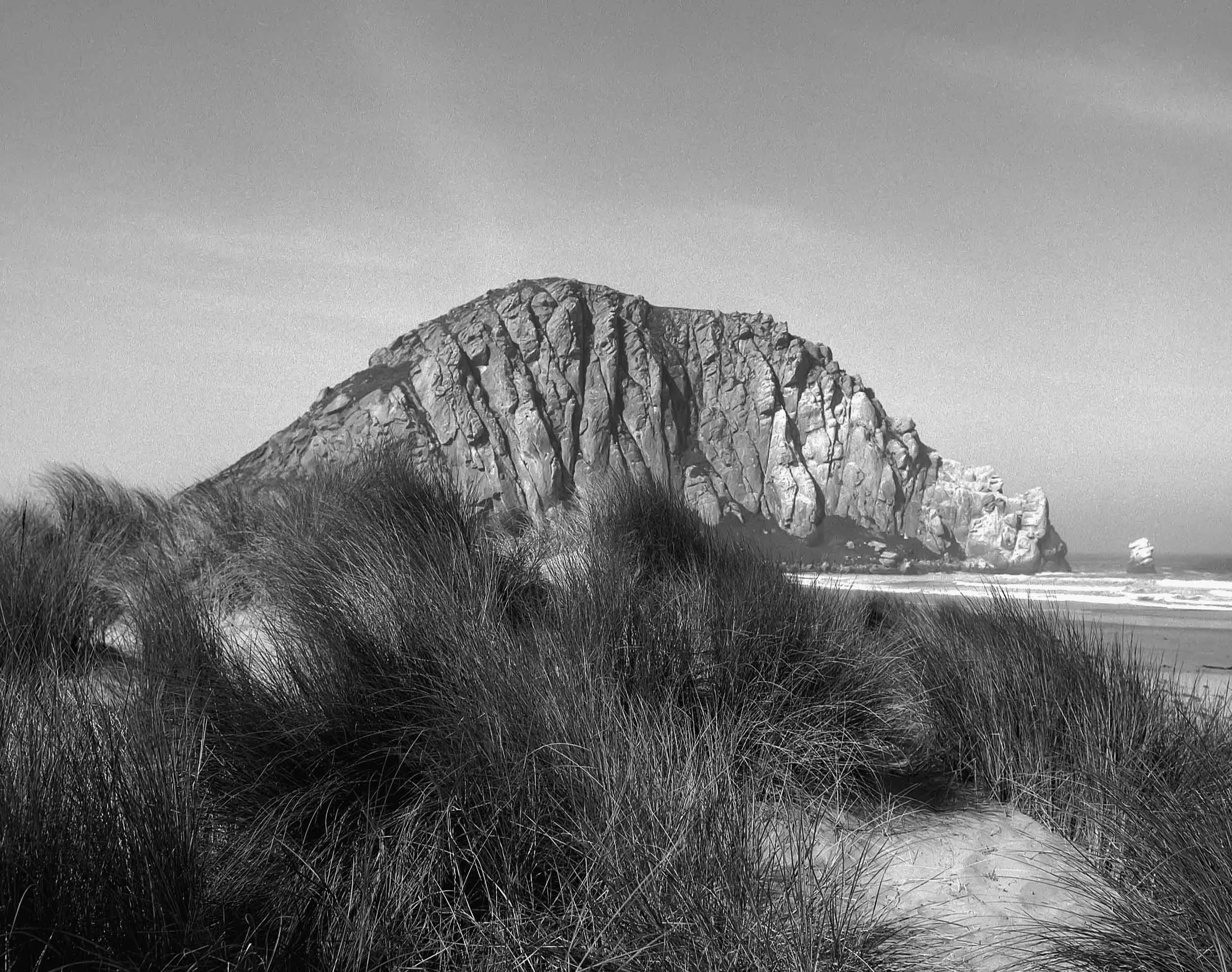 Black and white photograph of a large rocky mountain with sandy beach and grassy dunes in the foreground, under a clear sky.