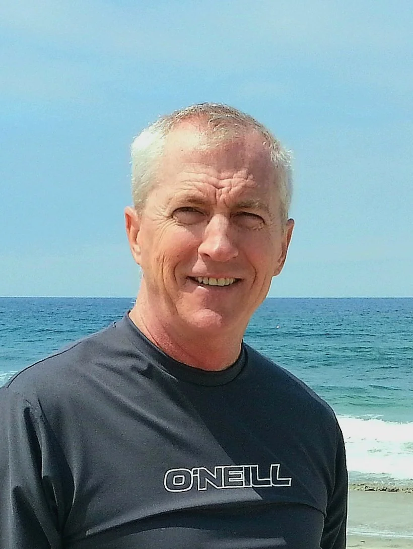 A smiling middle-aged man with short gray hair standing on a beach with the ocean and blue sky in the background, wearing a black O'Neill shirt.