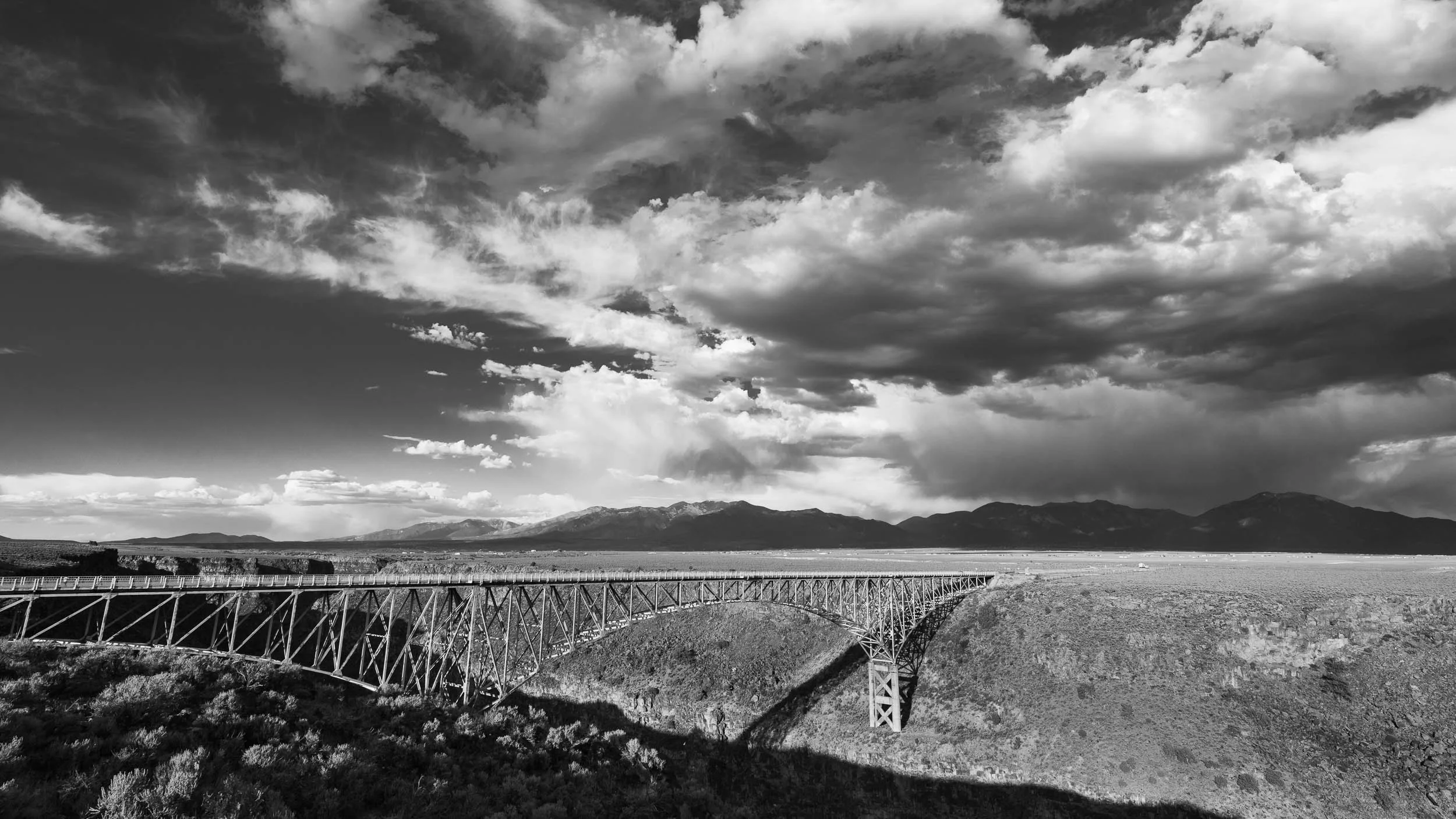Black and white photo of a large bridge spanning a canyon with mountains in the background and a partly cloudy sky.