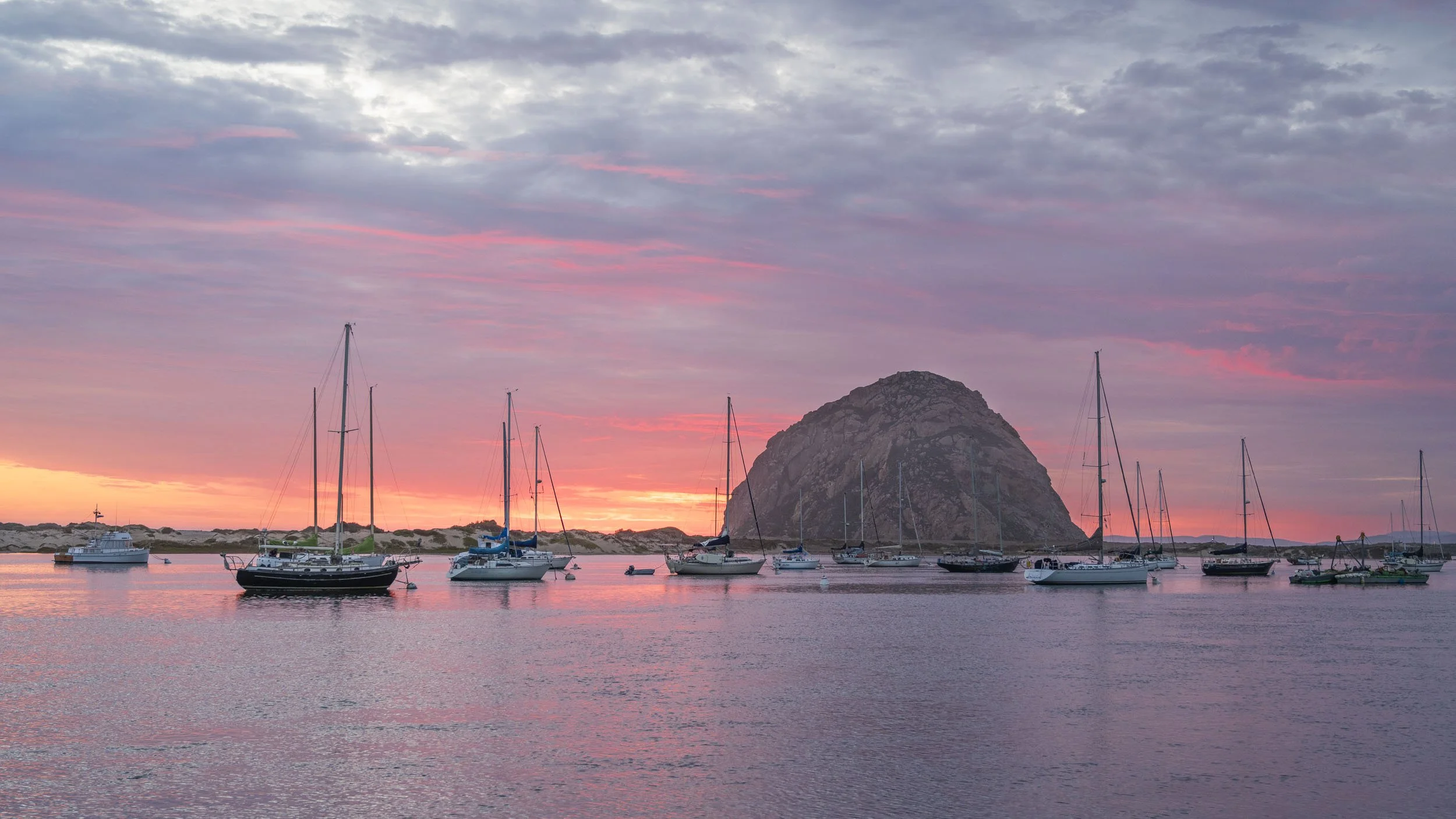 Sailboats anchored in a calm harbor during sunset with a large rock formation and a colorful sky with pink and purple clouds.