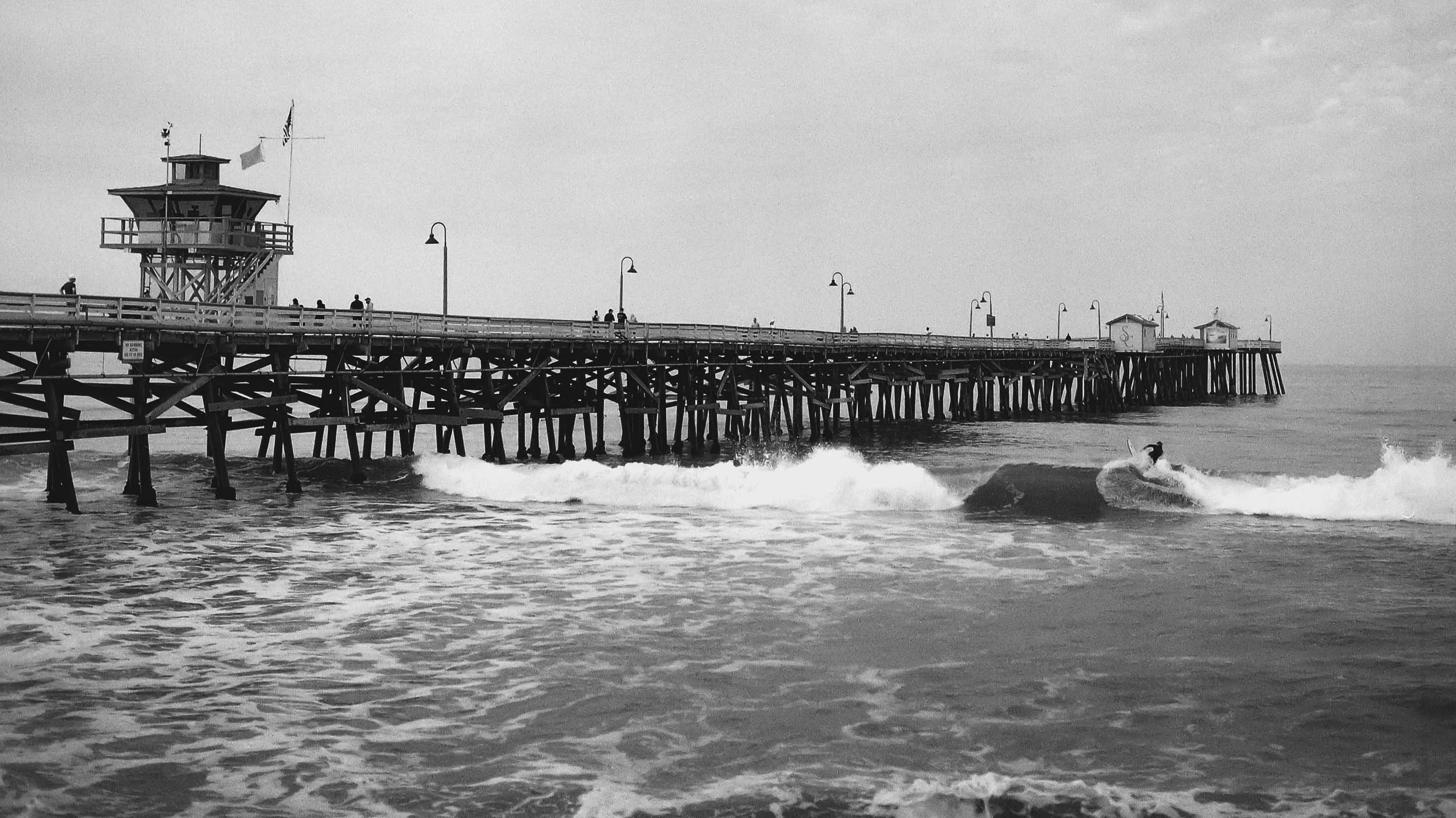 A black-and-white photo of a pier extending into the ocean, with a tower on the pier, lamp posts along the walkway, and people walking. A surfer is riding a wave in the ocean near the pier.
