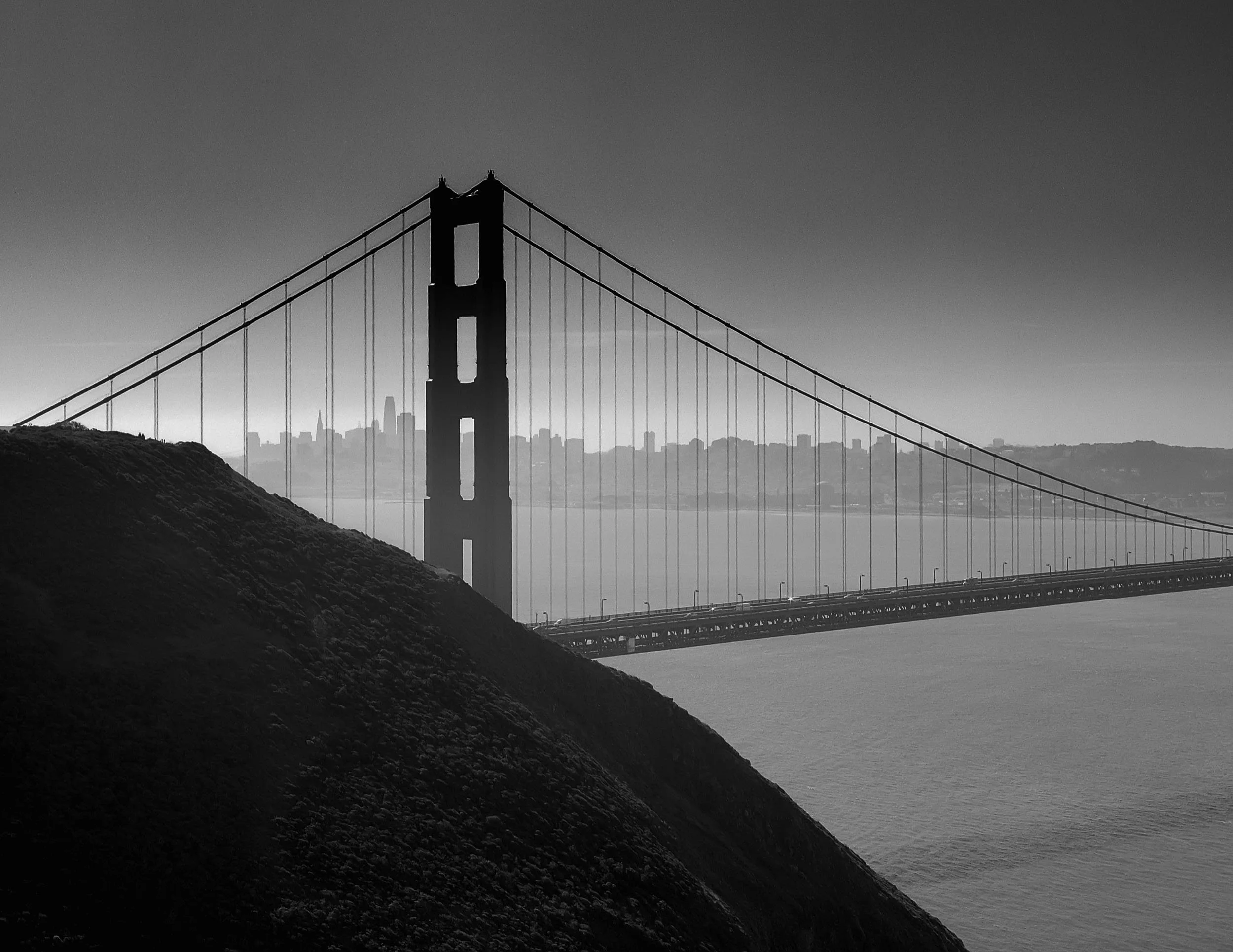 Black and white photo of the Golden Gate Bridge with foggy city skyline in the background, viewed from an elevated perspective over water, with dark rocky shoreline in the foreground.