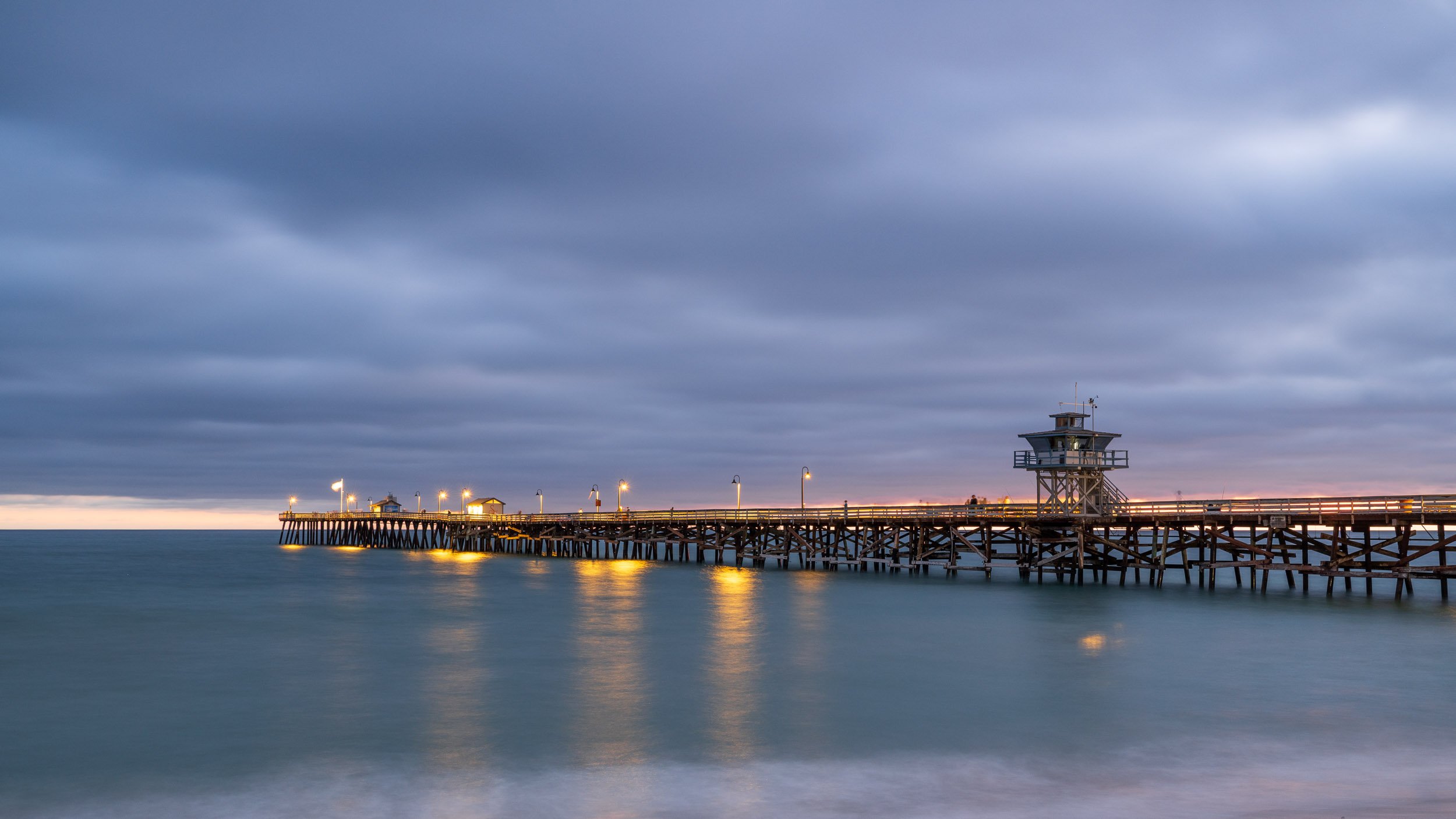A wooden pier extending into the ocean at dusk, with a small watchtower at the end and streetlights illuminating the walkway. The sky is overcast with dark clouds.