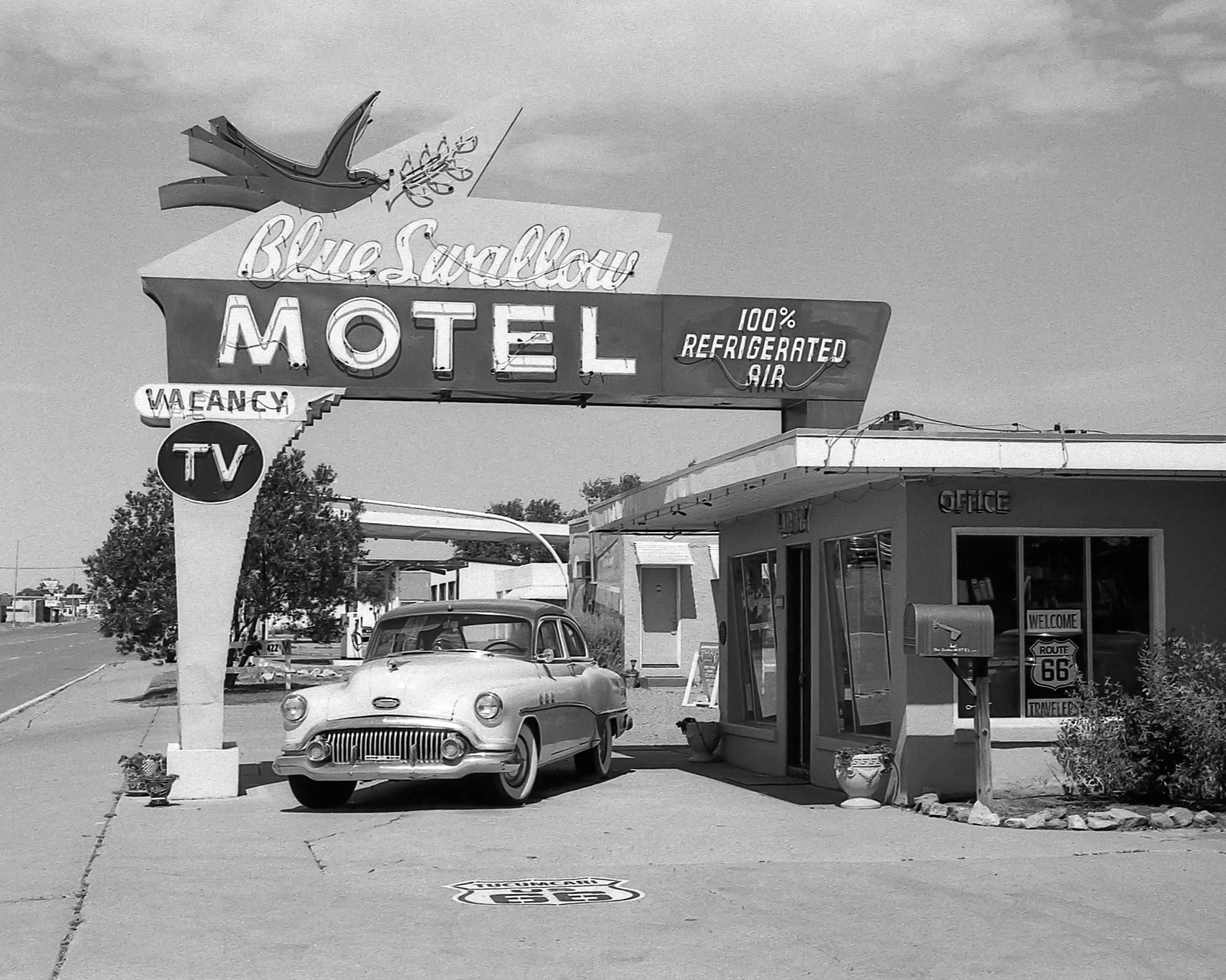 Black and white photo of a vintage car parked in front of a motel with a neon sign that reads 'Blue Swallow Motel 100% Refrigerated Air'. 