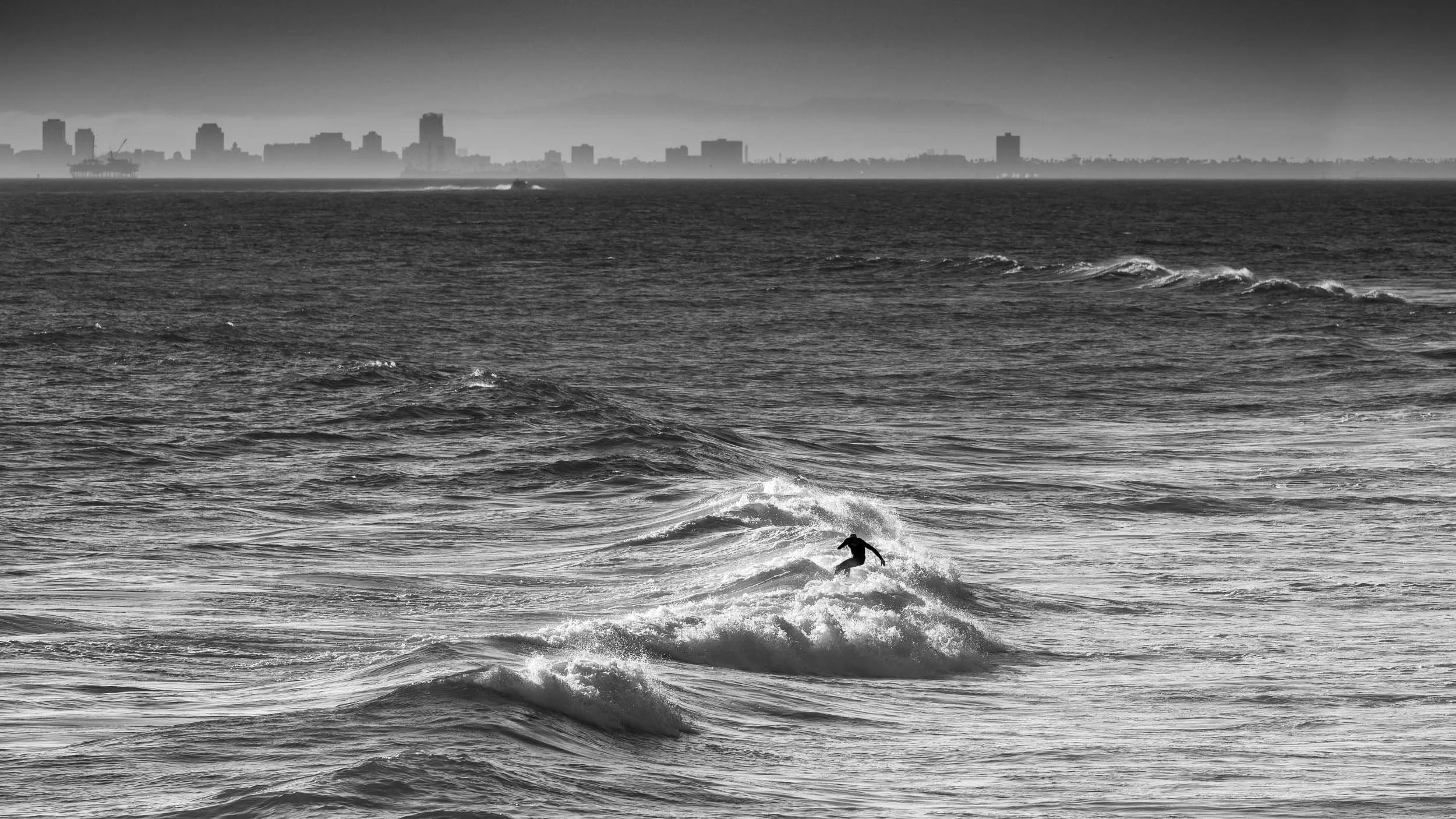 A person surfing on a wave in the ocean with a city skyline in the background in black and white.