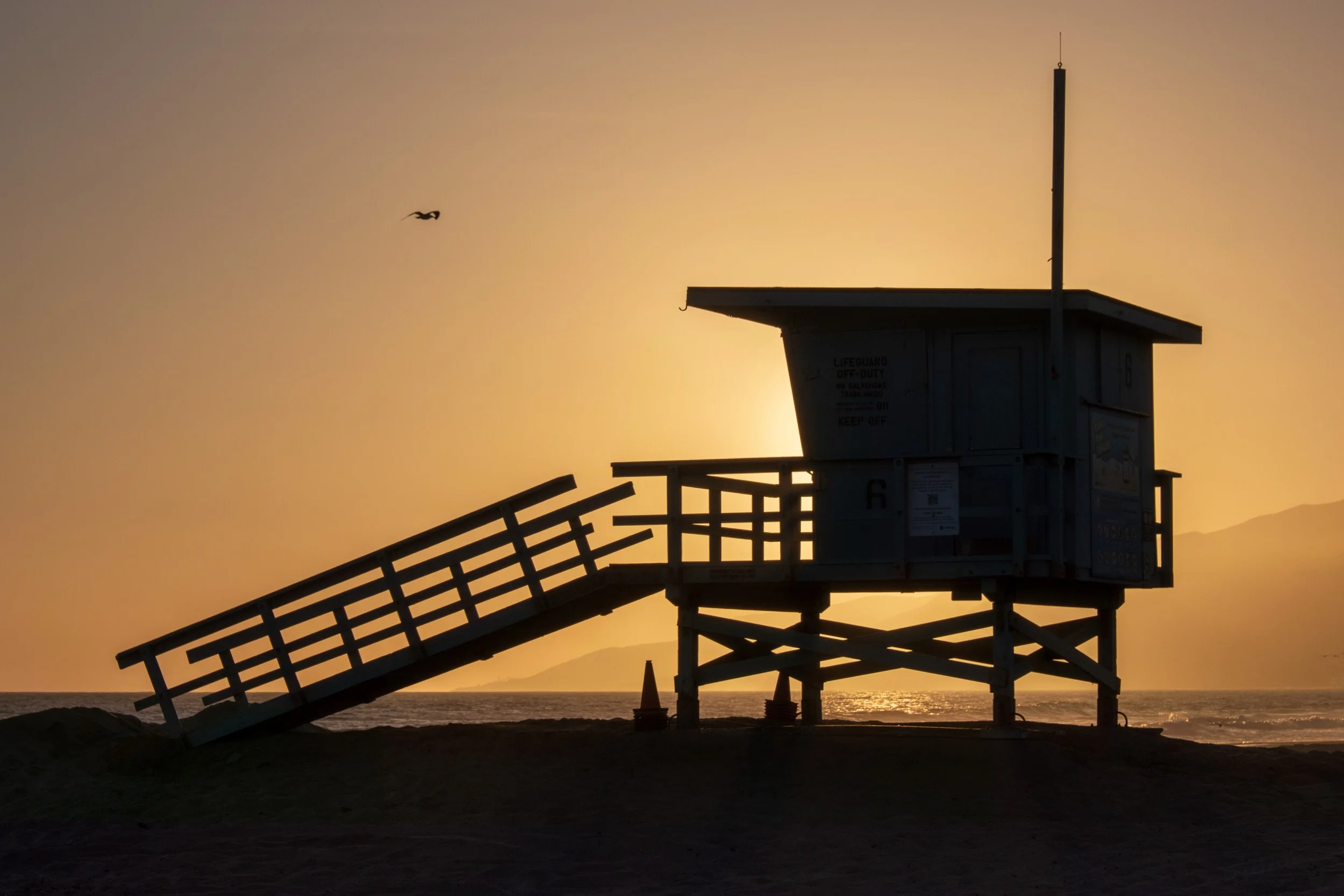 Silhouette of a lifeguard tower with a ramp, set against a sunset at the beach, with the ocean and mountains in the background.