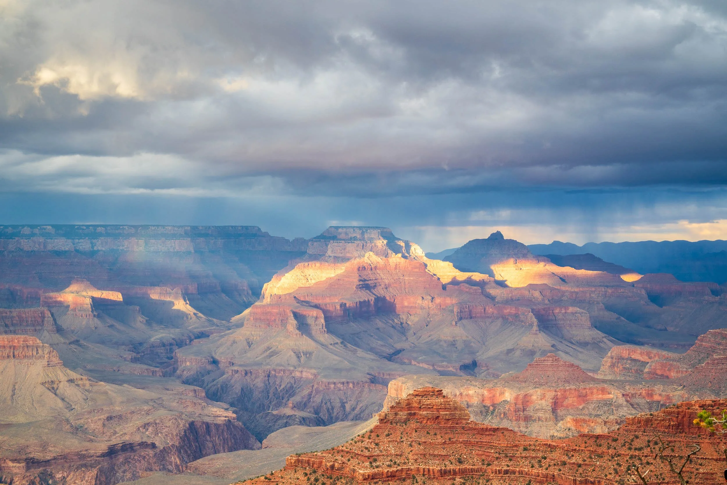 Panoramic view of the Grand Canyon under a cloudy sky, with layers of canyon walls illuminated by sunlight.