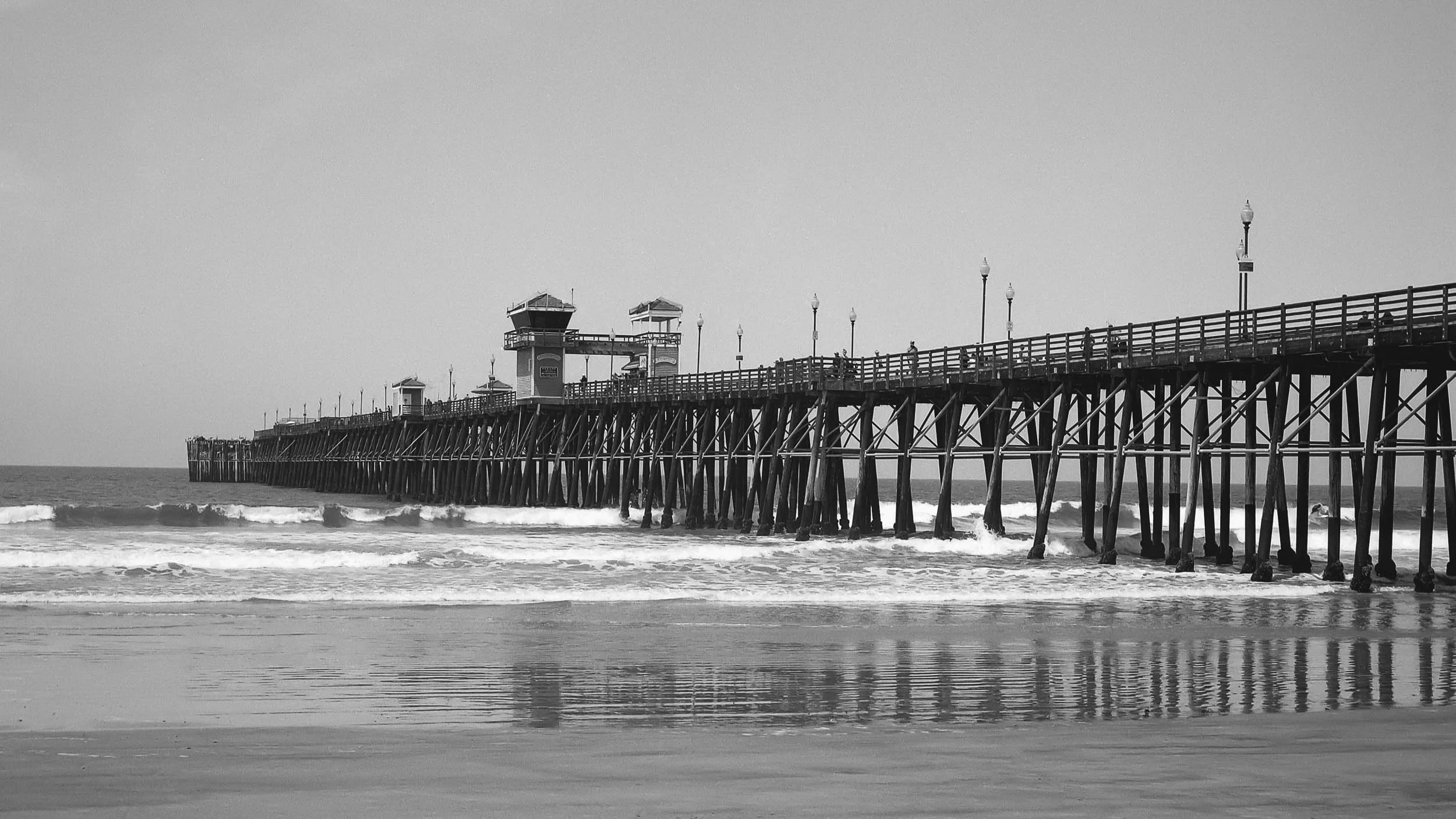 A black-and-white photo of a pier extending into the ocean with small waves crashing below, and several lampposts lining the pier. There are a few people walking on the pier, and the sky appears clear.