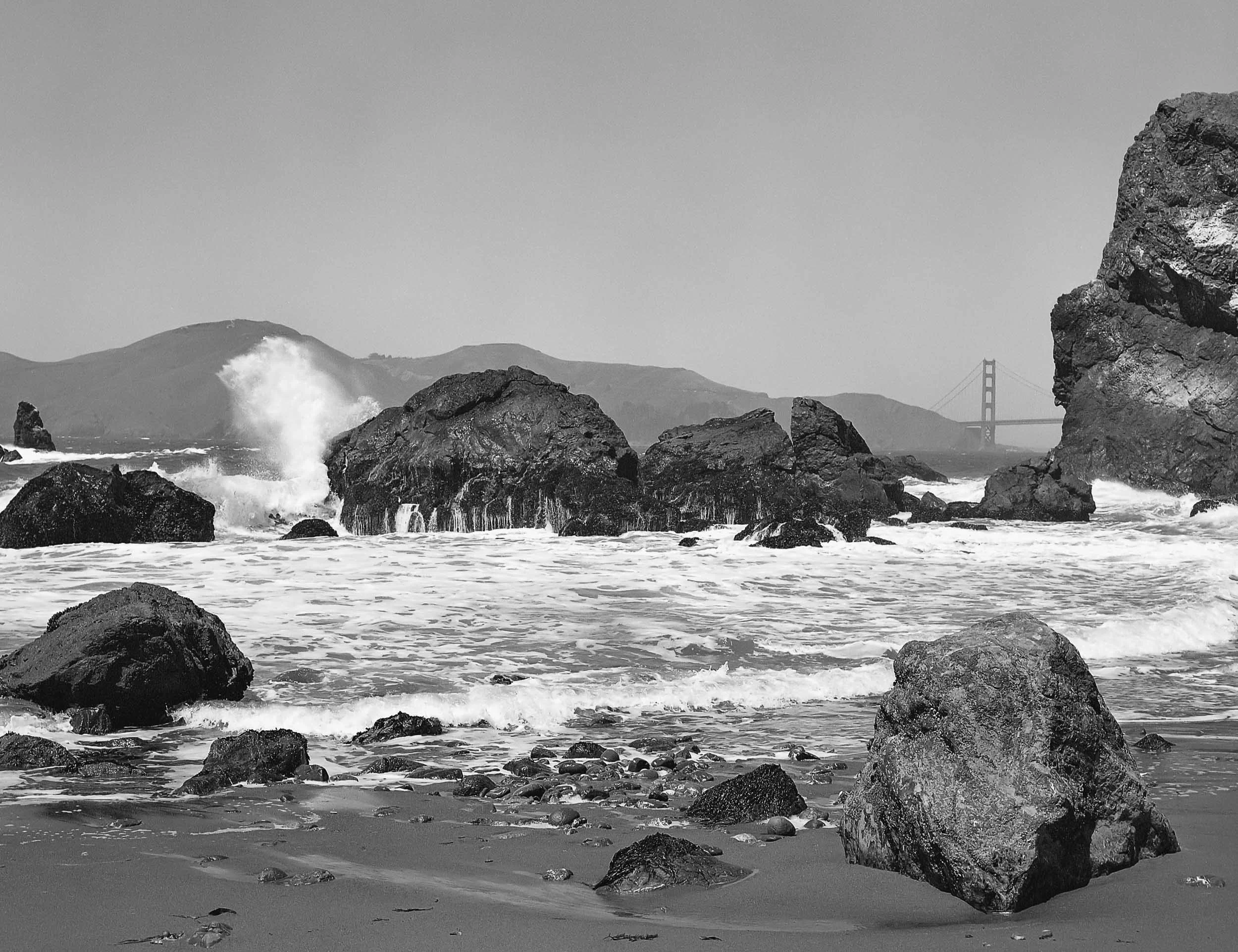 A black and white photo of a rocky shoreline with waves crashing against rocks, with hills and a bridge in the distance.