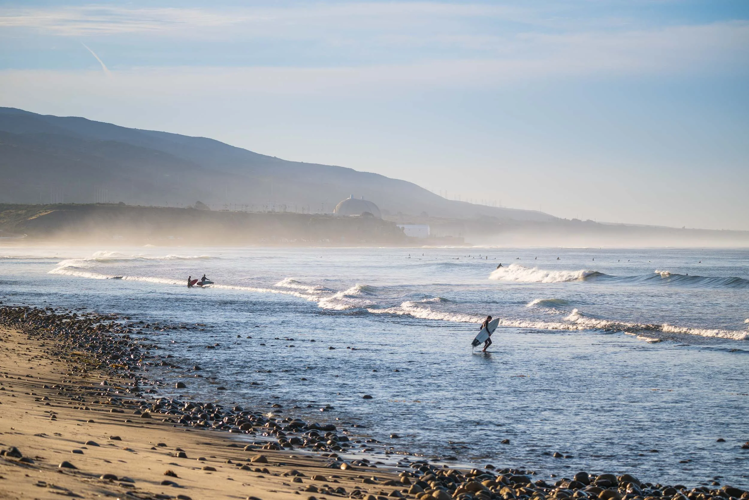 A scenic beach with surfers carrying surfboards, waves crashing on the shore, and a misty coastline with mountains in the background under a partly cloudy sky.