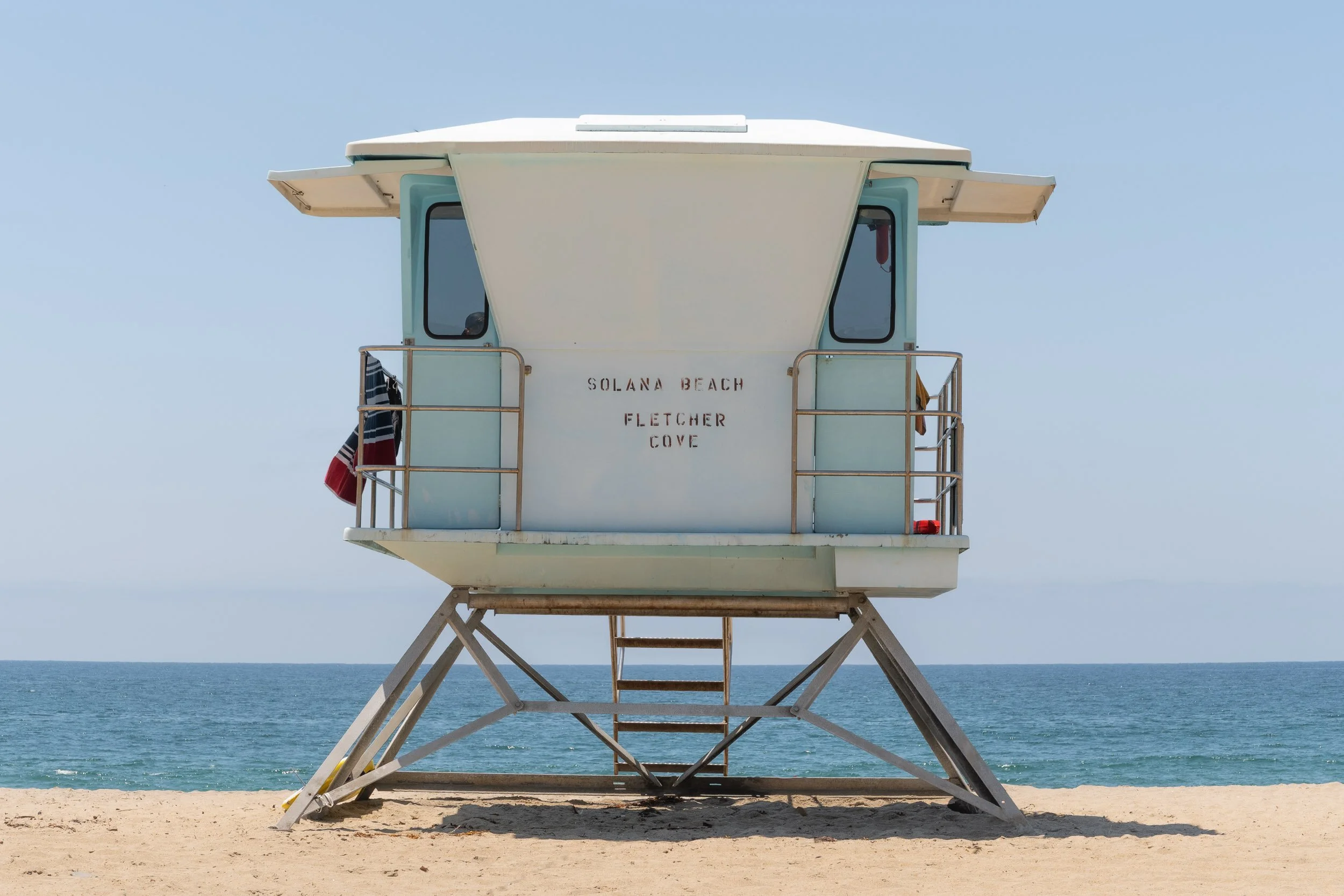 A lifeguard tower labeled "Solana Beach Fletcher Cove" stands on the sandy beach with the ocean and clear blue sky in the background.