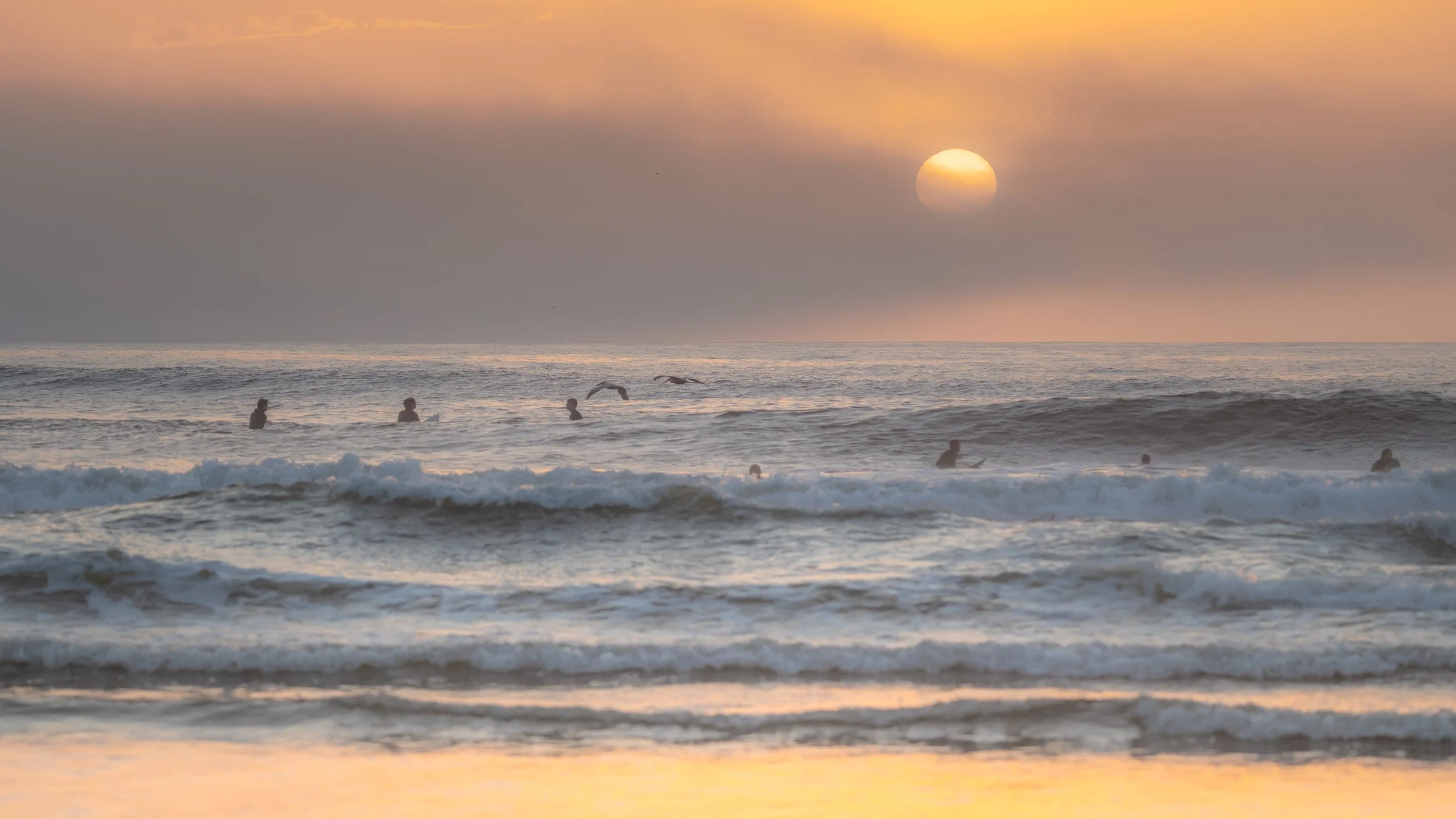 People swimming and surfing in the ocean during sunset with a cloudy sky and the sun low on the horizon.