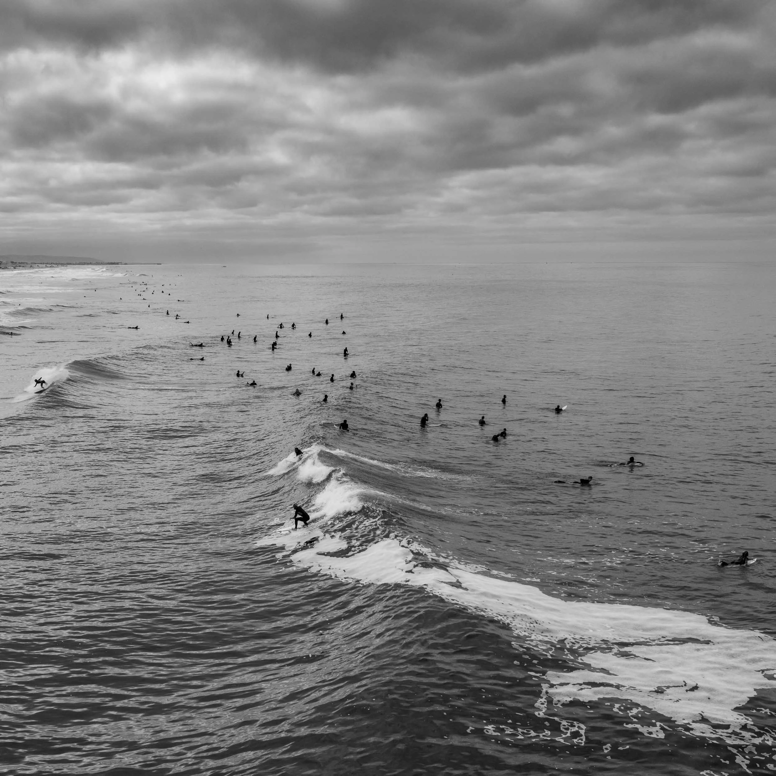 Black and white photo of an ocean with many surfers in the water waiting for waves, and a cloudy sky overhead.