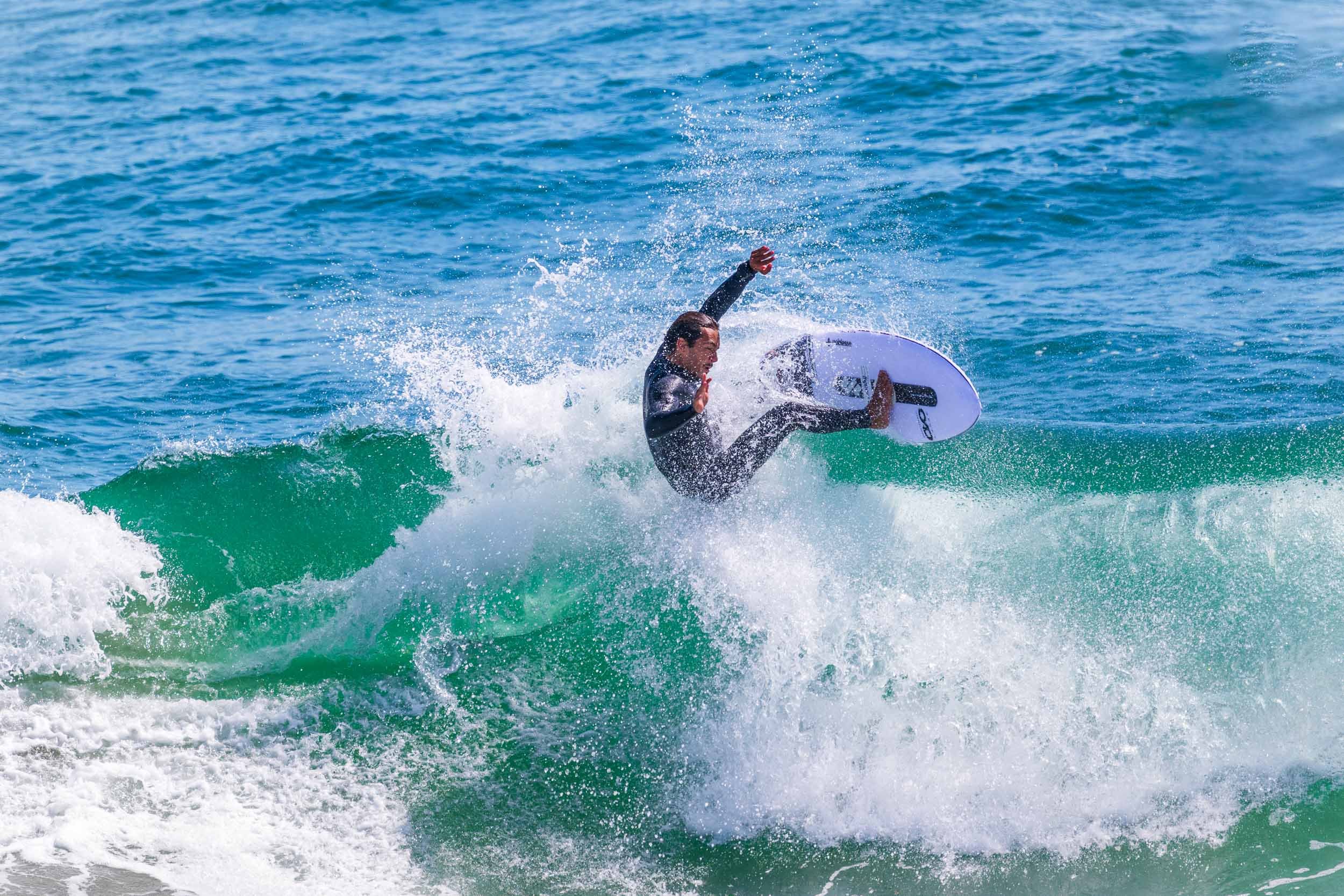 A person wearing a wetsuit surfing on a white surfboard with a black logo on a blue wave in the ocean, surrounded by splashing water.