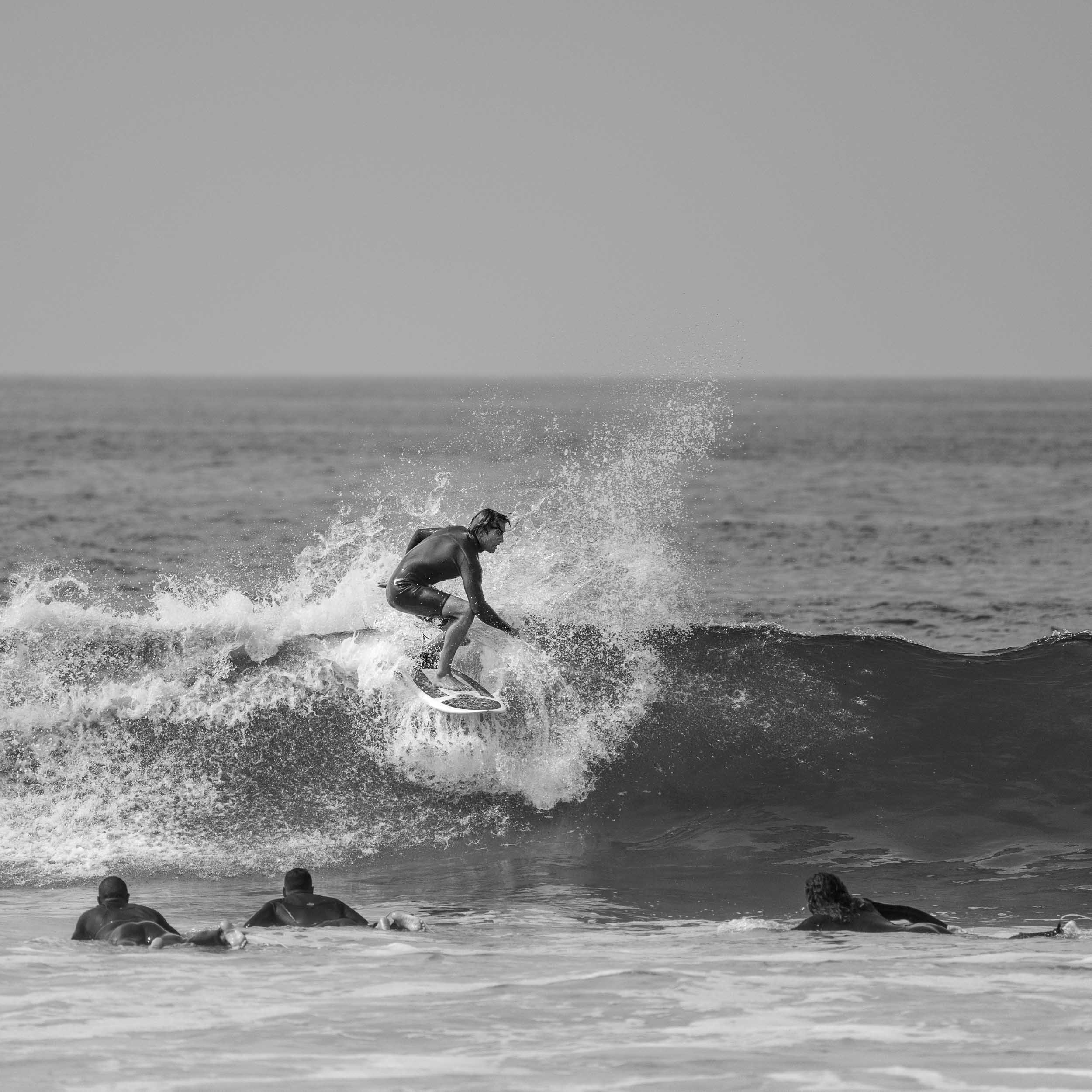 A group of surfers watching a person ride a wave in the ocean on a sunny day.