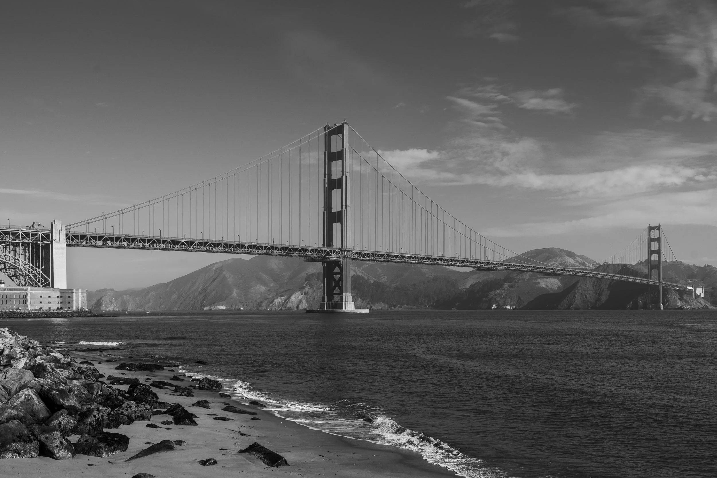 Black and white photo of the Golden Gate Bridge in San Francisco, with a rocky shoreline and water in the foreground, and mountains in the background.