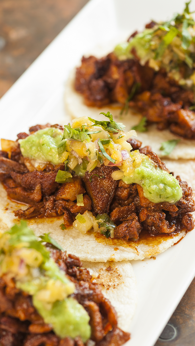 Close-up of three tacos with seasoned beef, guacamole, and pico de gallo on soft corn tortillas, served on a white plate.