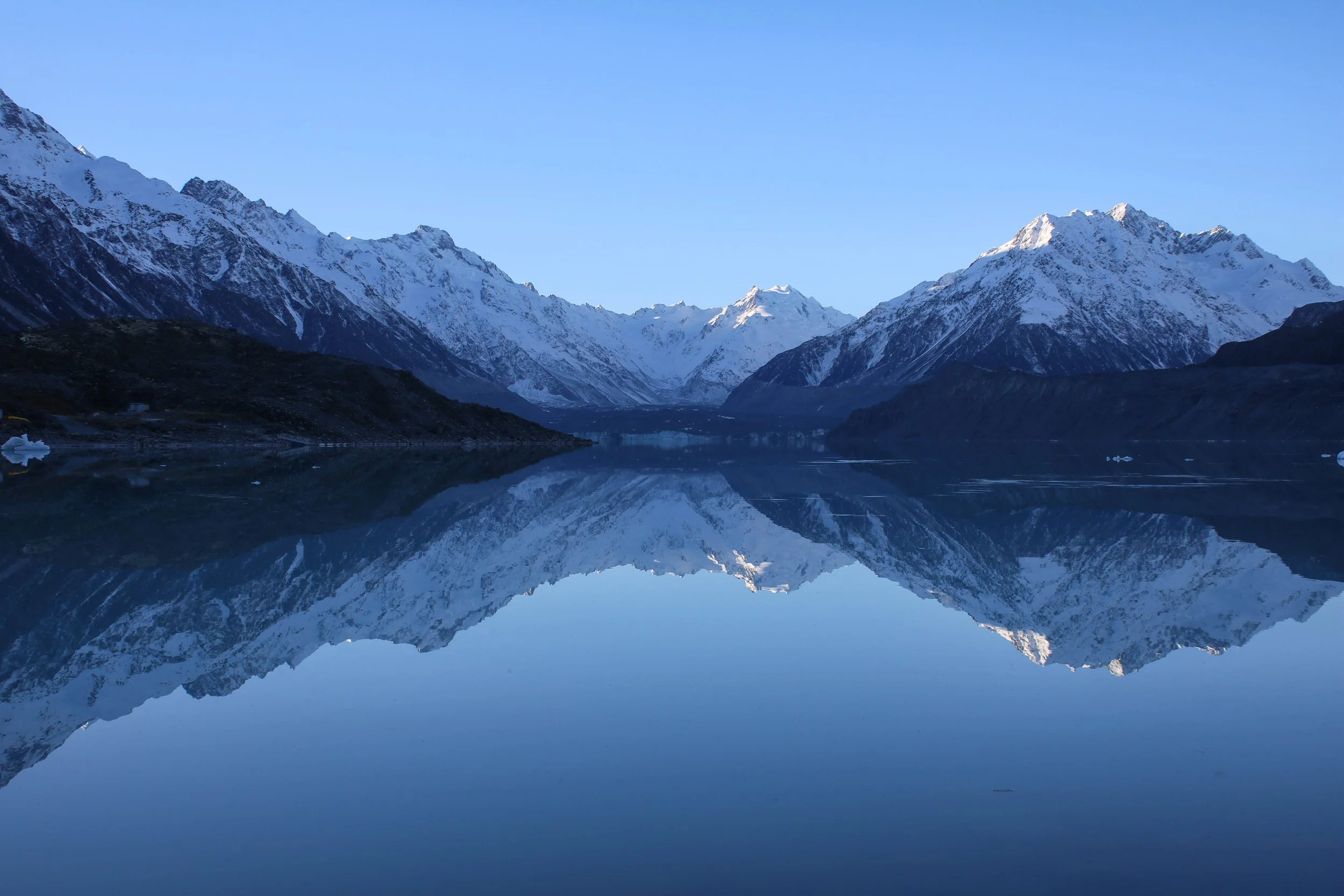 Snow-covered mountains reflected in a calm lake under a clear blue sky at Mt Cook Glacier