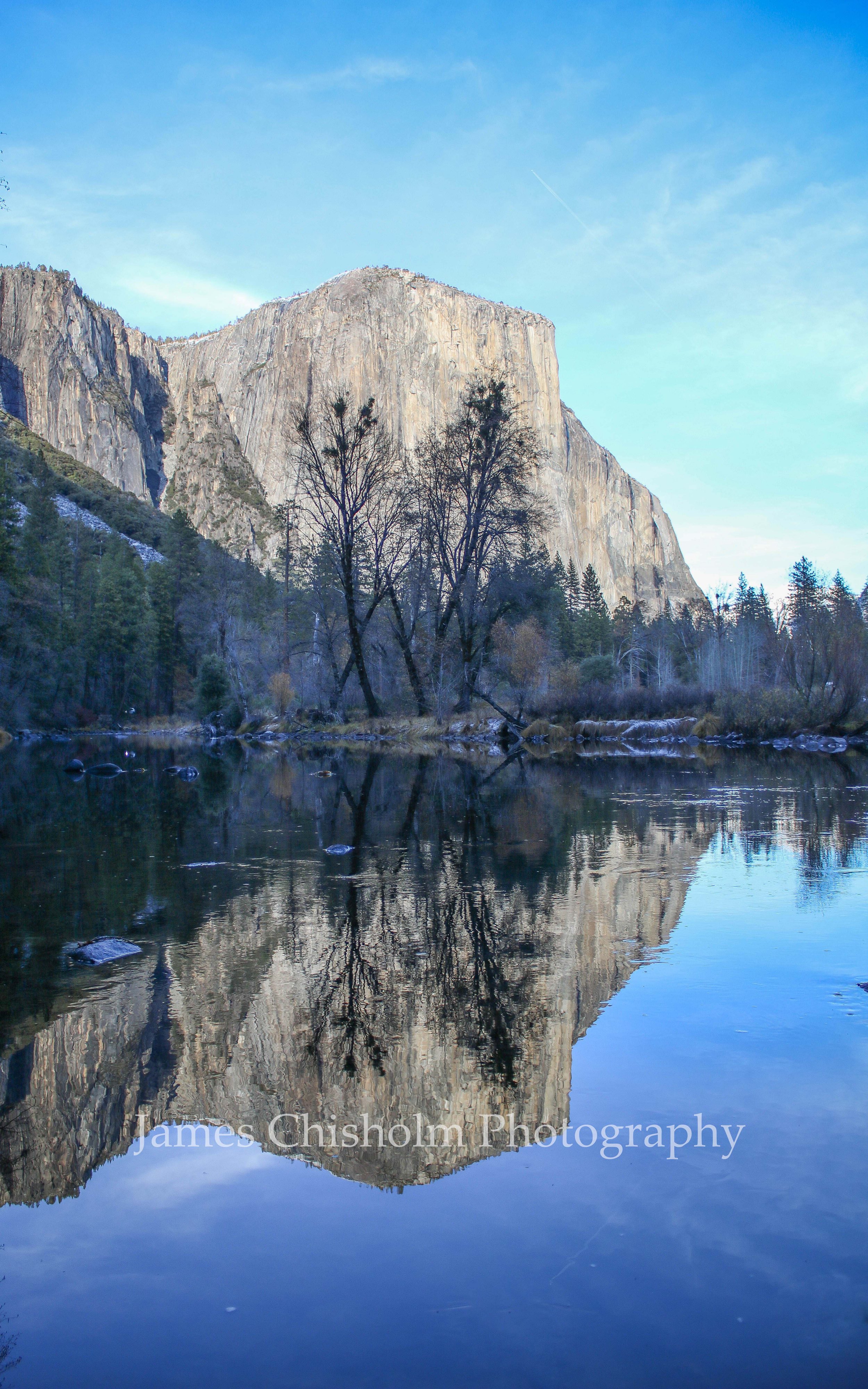 Reflected view of a granite cliff and trees along a river under a blue sky