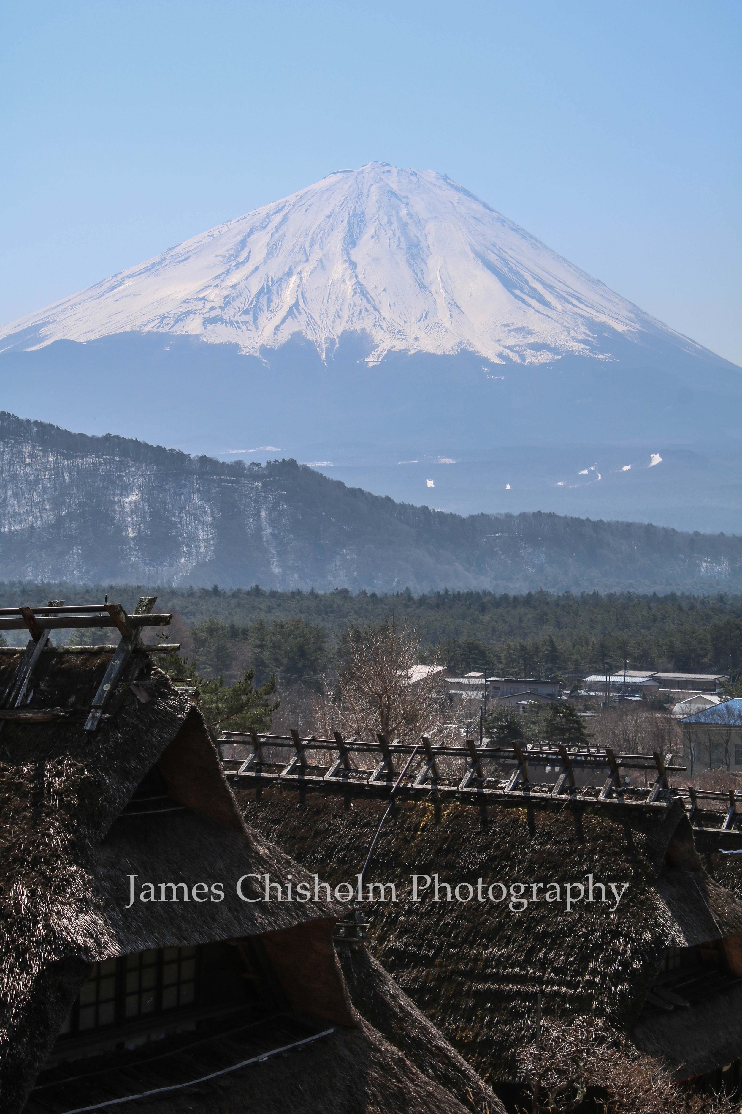 Snow-capped Mount Fuji over a rural village with traditional thatched roofs and trees, under a clear blue sky.