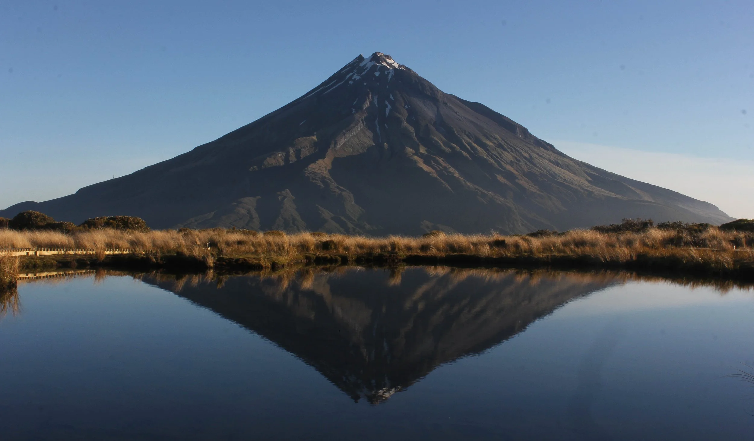 A snow-capped volcano in New Zealand, reflected in a calm body of water with grassy edges, under a clear blue sky at the Pouakai Tarns, Taranaki.
