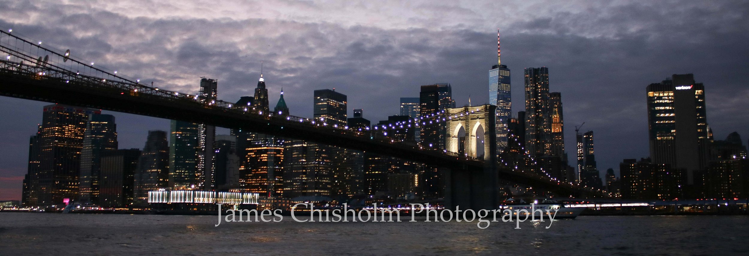 Nighttime view of the New York City skyline with Brooklyn Bridge in the foreground, illuminated skyscrapers in Manhattan, and cloudy sky.