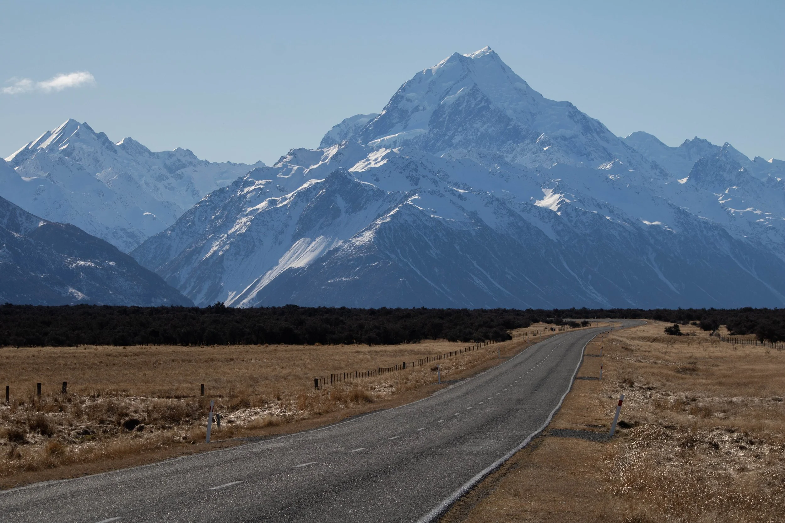 A winding rural road leading towards snow-capped mountains in the distance, with open fields and a fence in the foreground on the way to Mt Cook National Park.