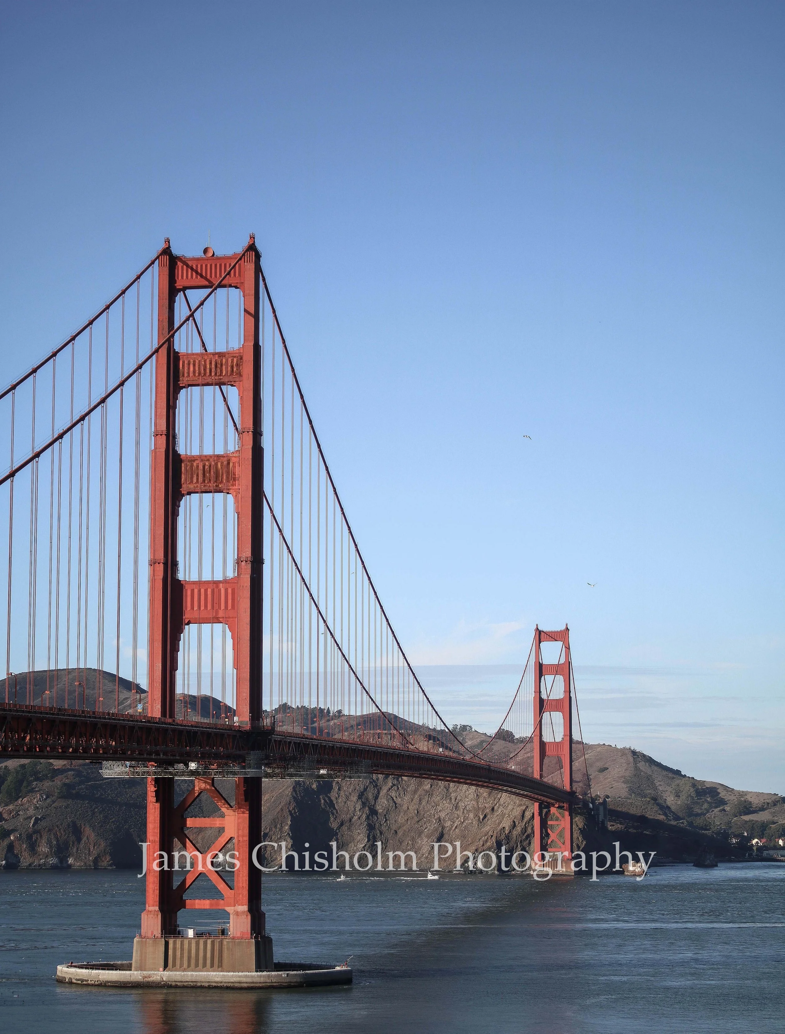 View of the Golden Gate Bridge in San Francisco, California, with the bridge's iconic reddish-orange towers and suspension cables visible, set against a blue sky with some clouds and hills in the background.