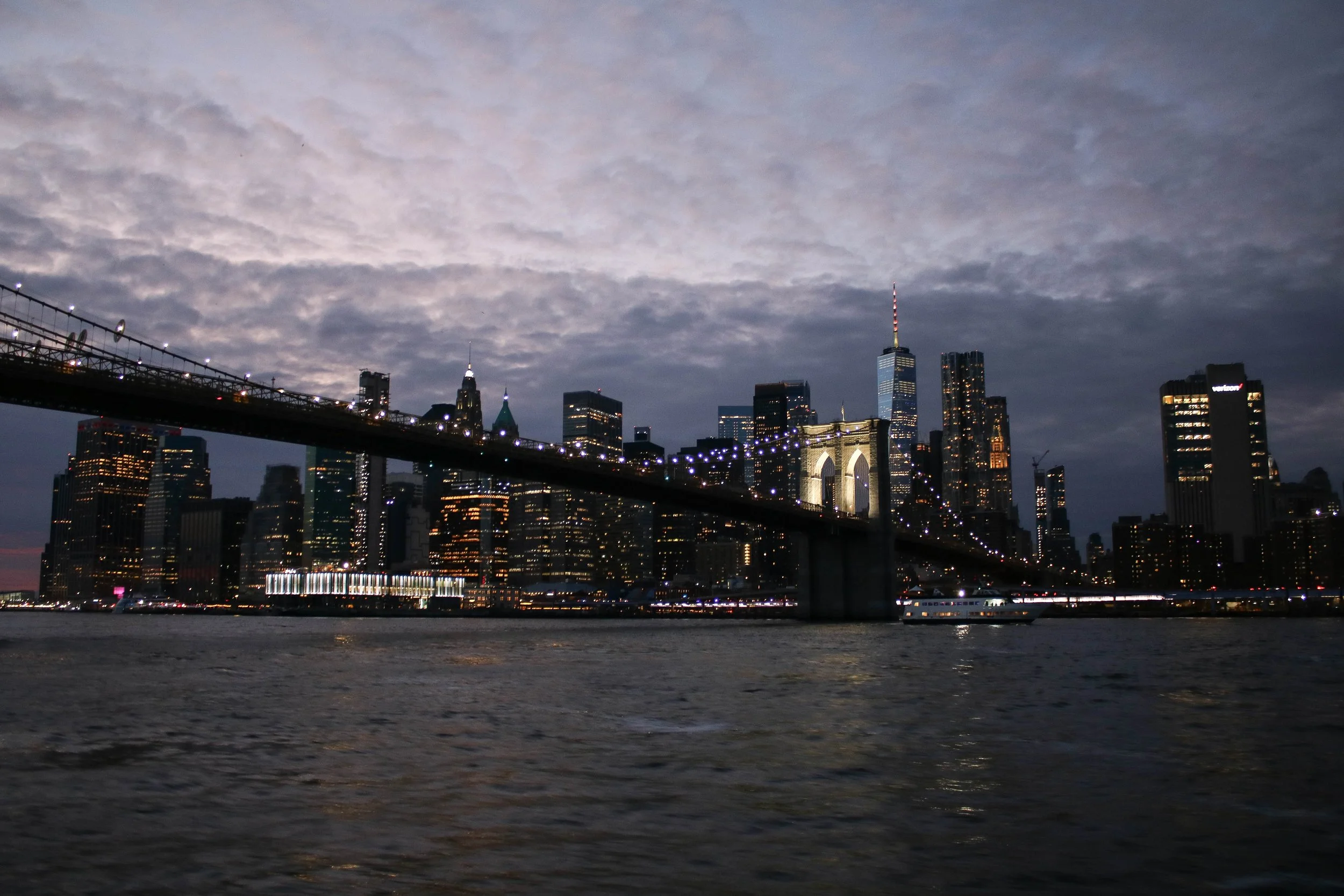 Nighttime view of a city skyline with tall buildings, including the Empire State Building, and a bridge with lights over a body of water with reflections.