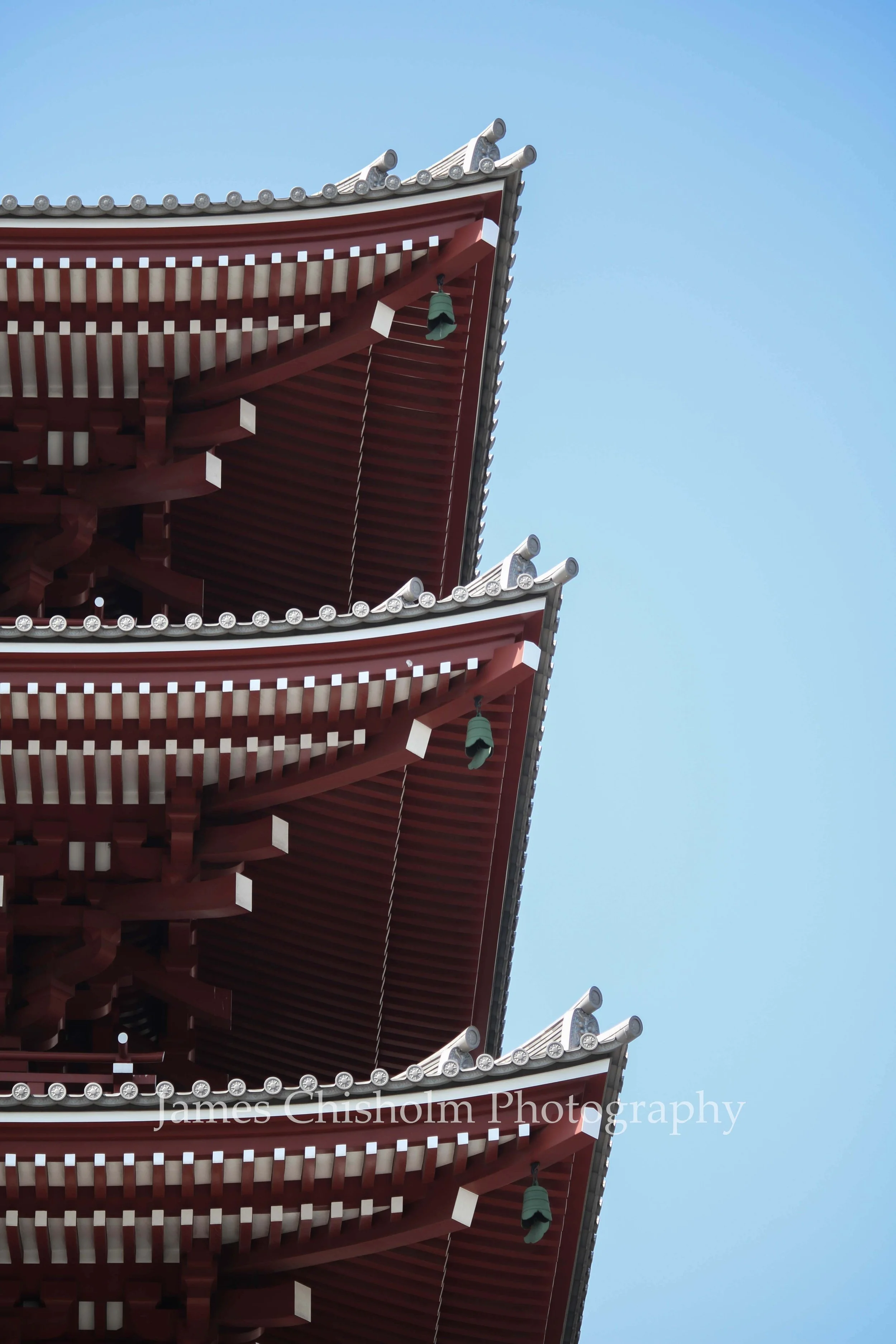 Top of a traditional Japanese pagoda with ornate roof details and hanging bells against a clear blue sky.