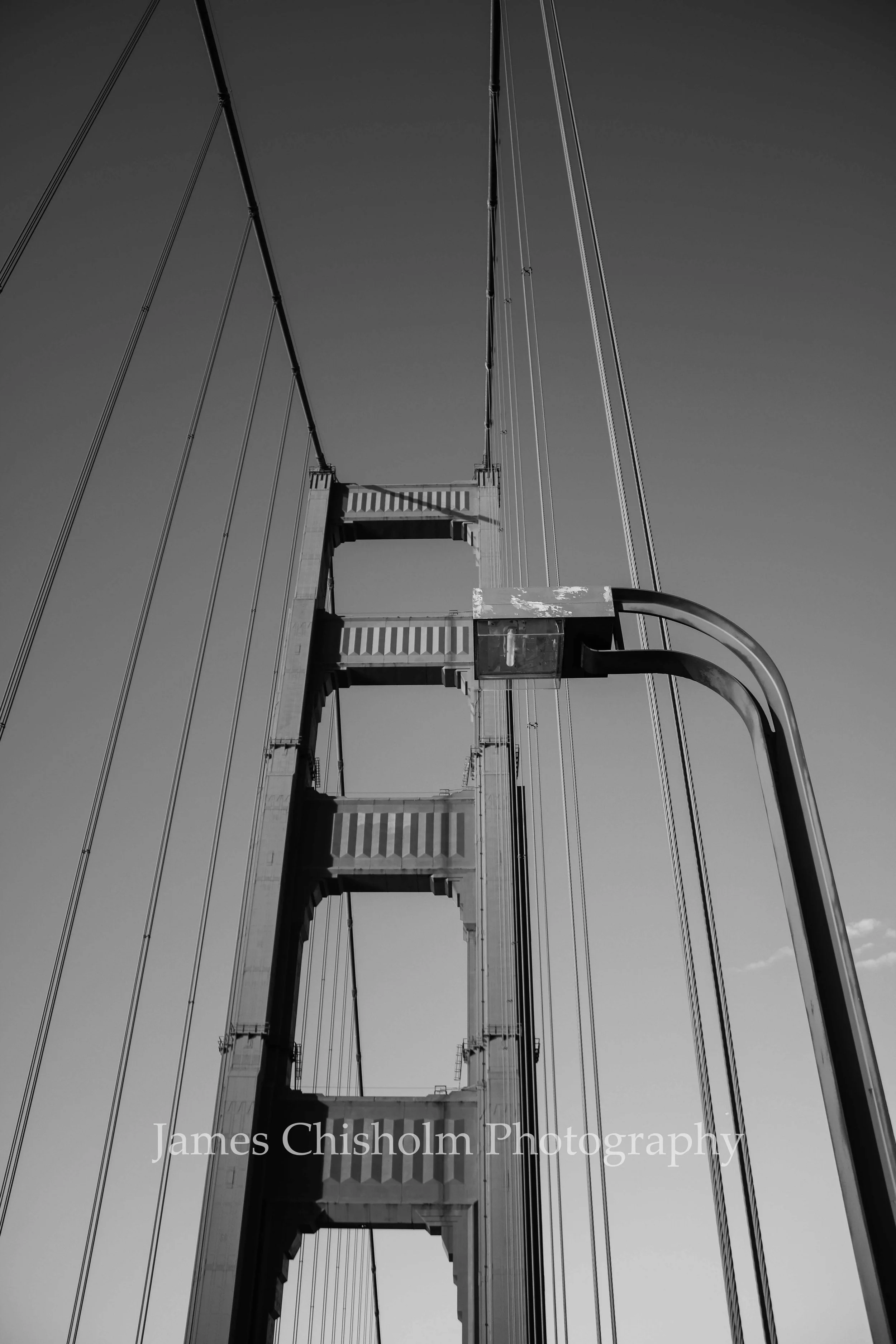 Black and white photo of the suspension bridge's tall towers, cables, and a roadway, with the sky in the background.