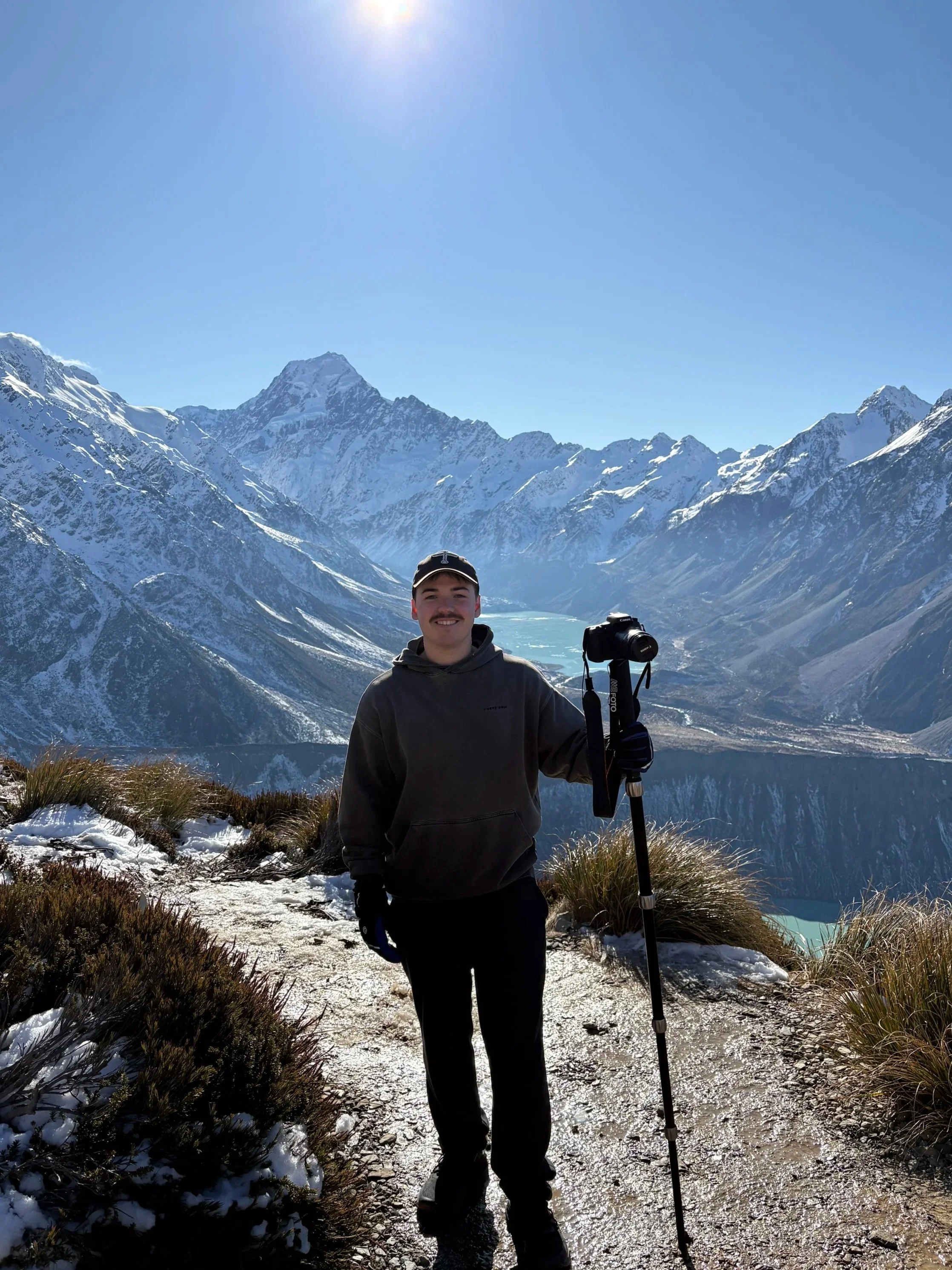 A young man standing on a snowy trail in a mountain landscape with snow-covered peaks, a lake, and a clear blue sky in the background, holding a camera on a tripod.