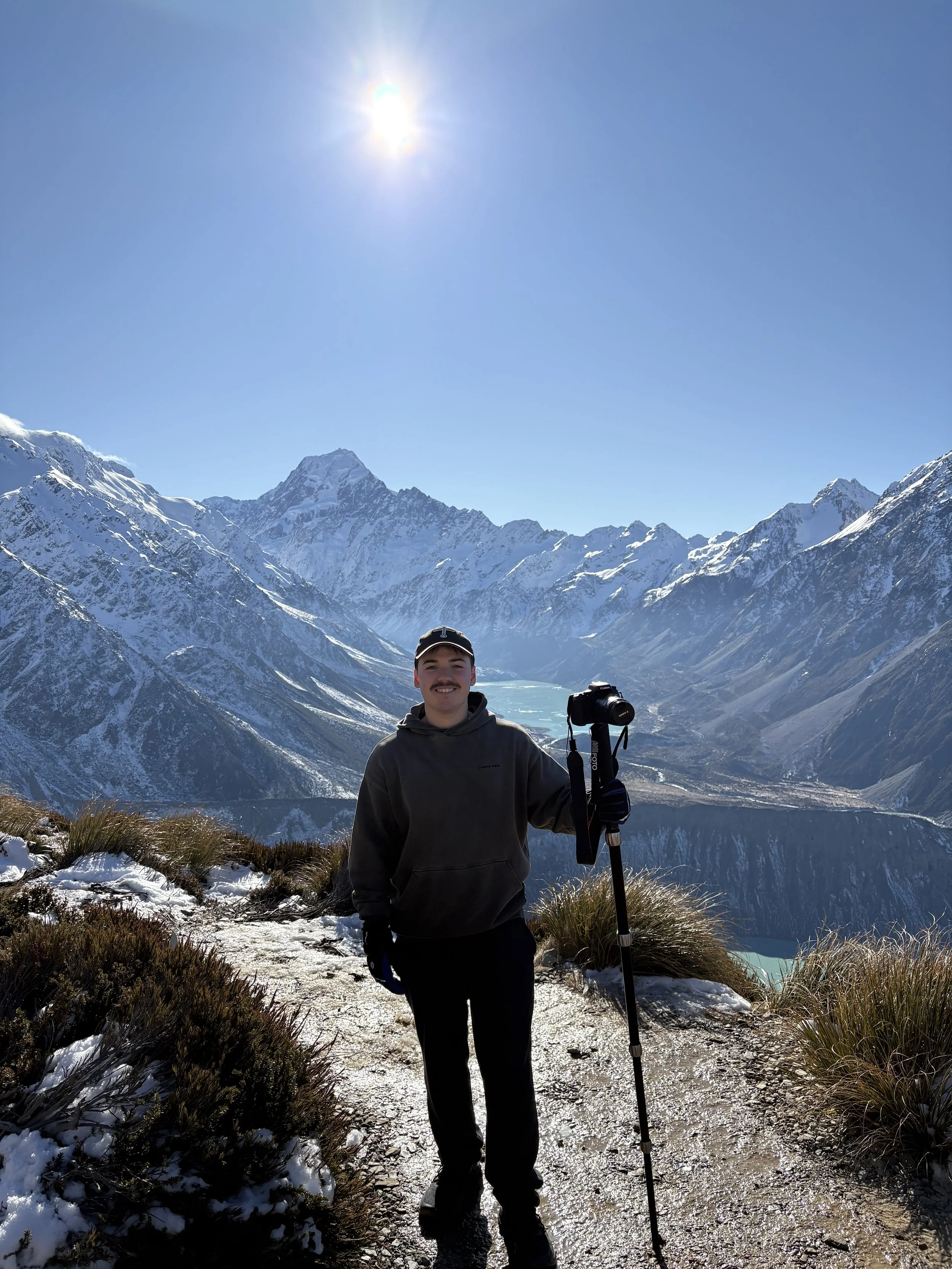 A man smiling in a black jacket and pants stands on a snow-covered mountain trail, holding a trekking pole, with snow-capped mountains and a bright sun in the clear blue sky in the background.