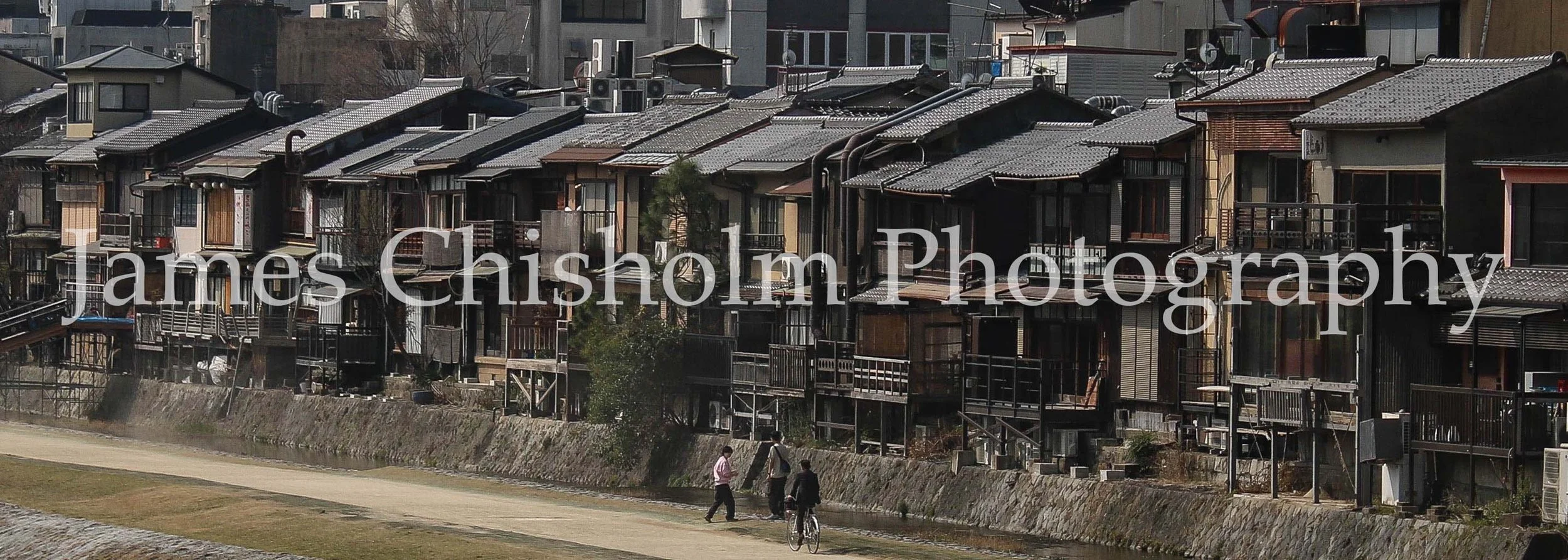 A row of traditional Japanese-style houses with tiled roofs along a canal, with three people walking and a person riding a bicycle on the path beside the canal.