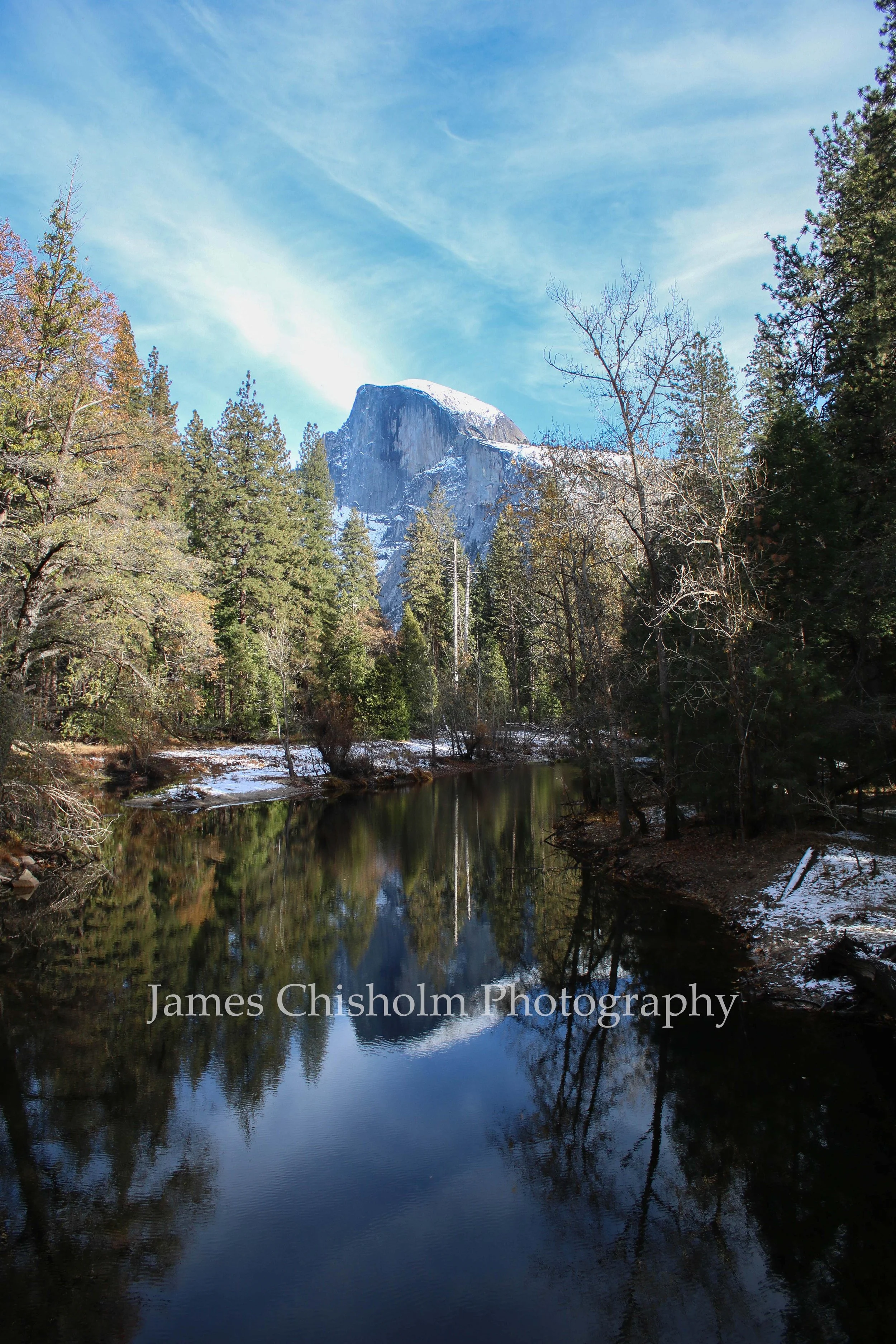 A tall, snow-covered granite mountain at Yosemite rises behind a framed forest and calm river reflecting trees and sky, under a blue sky with wispy clouds.