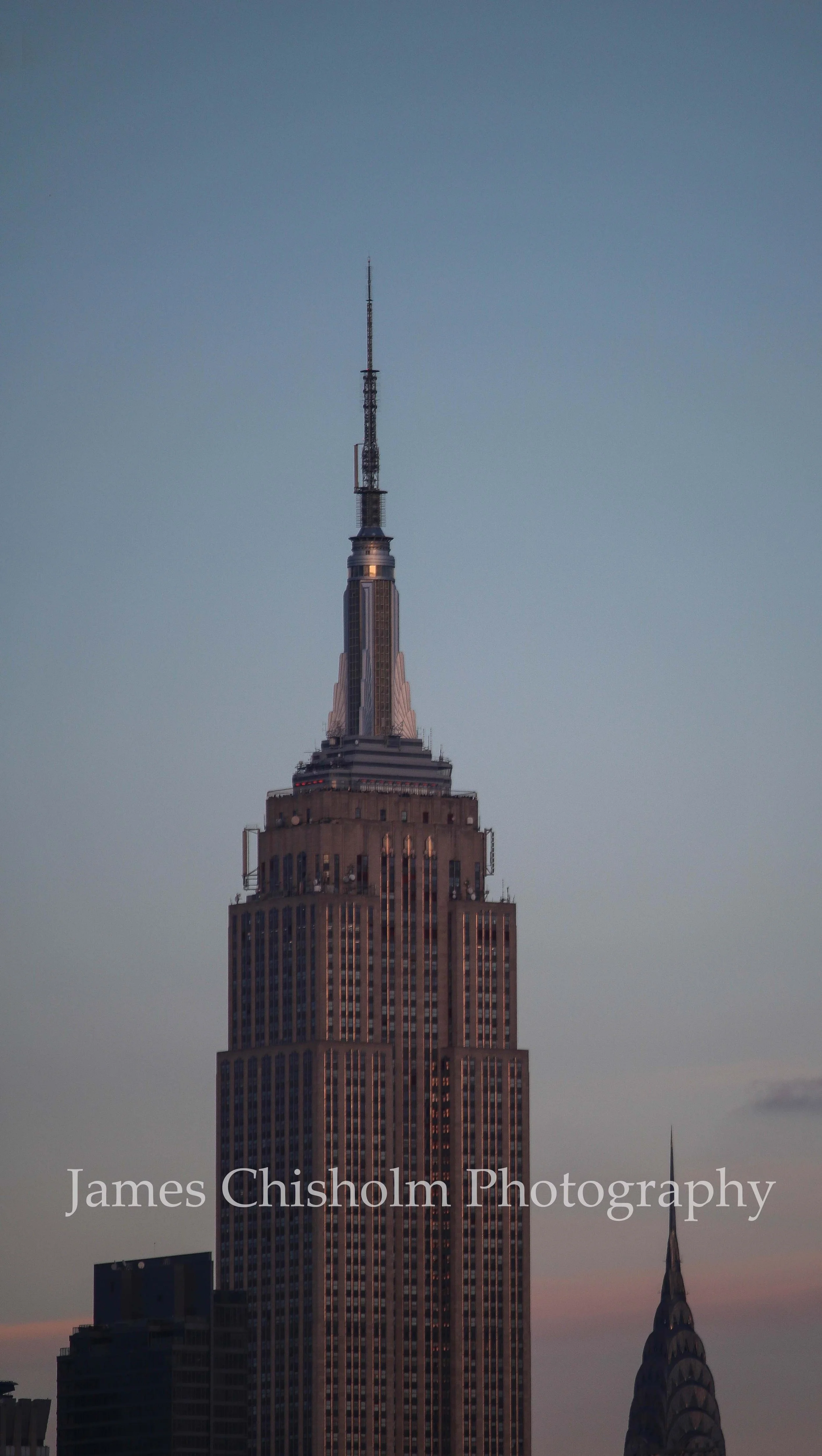 The Empire State Building at dusk with its illuminated top, and the spire of the Chrysler Building nearby, in New York City skyline.