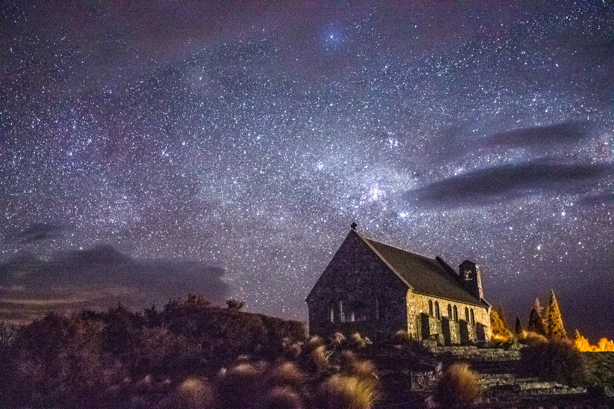 Night sky in New Zealand filled with countless stars and the Milky Way galaxy visible above a house with a steep roof, with some dark clouds and illuminated trees and landscape in the foreground.