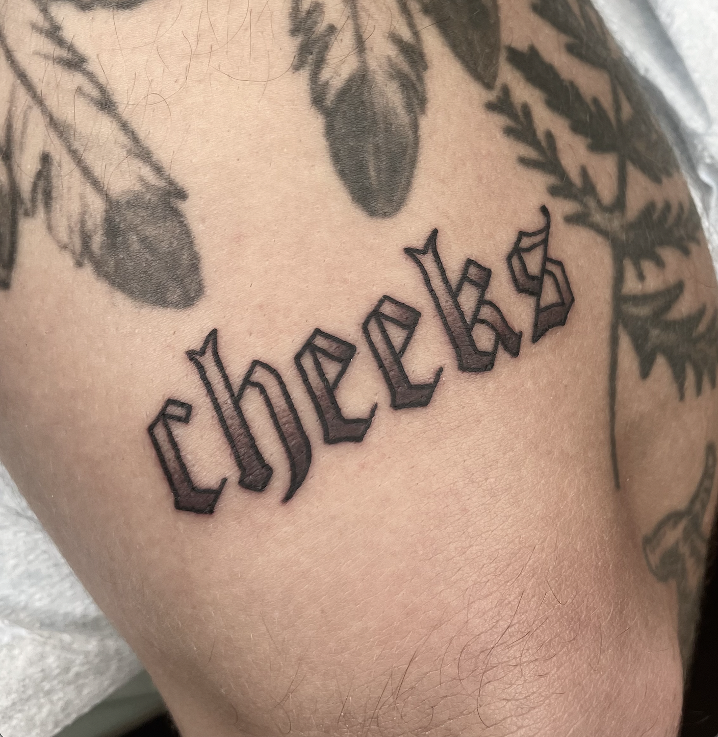 Close-up of a tattoo on skin with the word 'cheeks' in gothic-style font, surrounded by other tattoos including floral designs and dark shading.